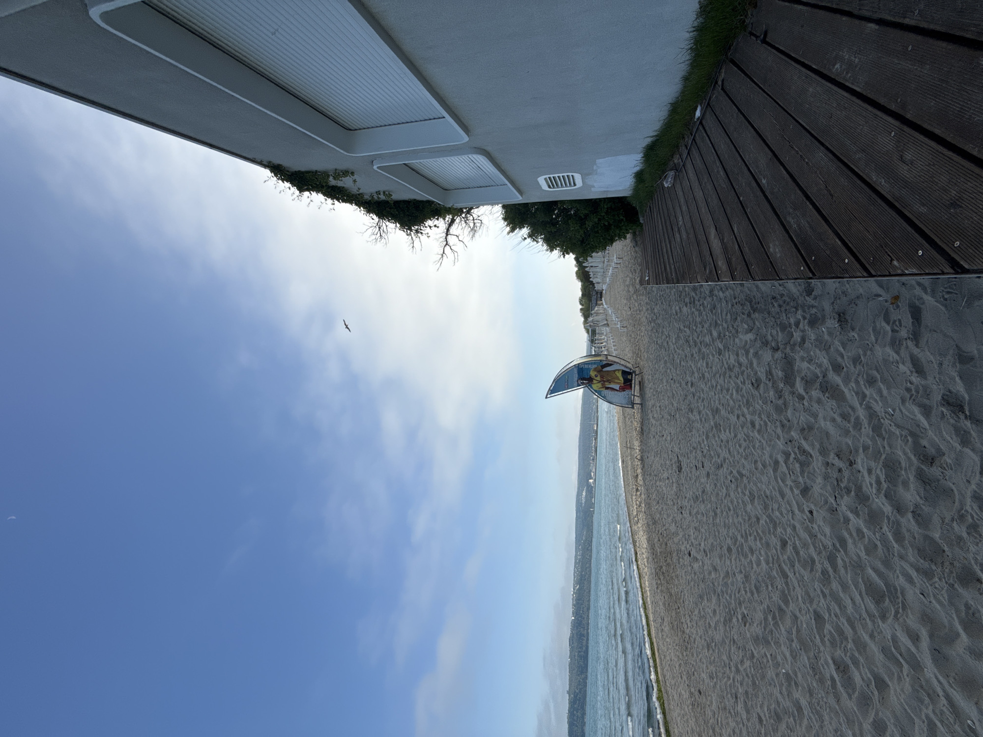 The image shows a beach scene in Varna, Bulgaria. A sandy beach is located next to the Black Sea. A wooden deck is on the right. A building is on the left. There is a sign on the beach. The sky is blue with some clouds. A bird is flying in the sky.