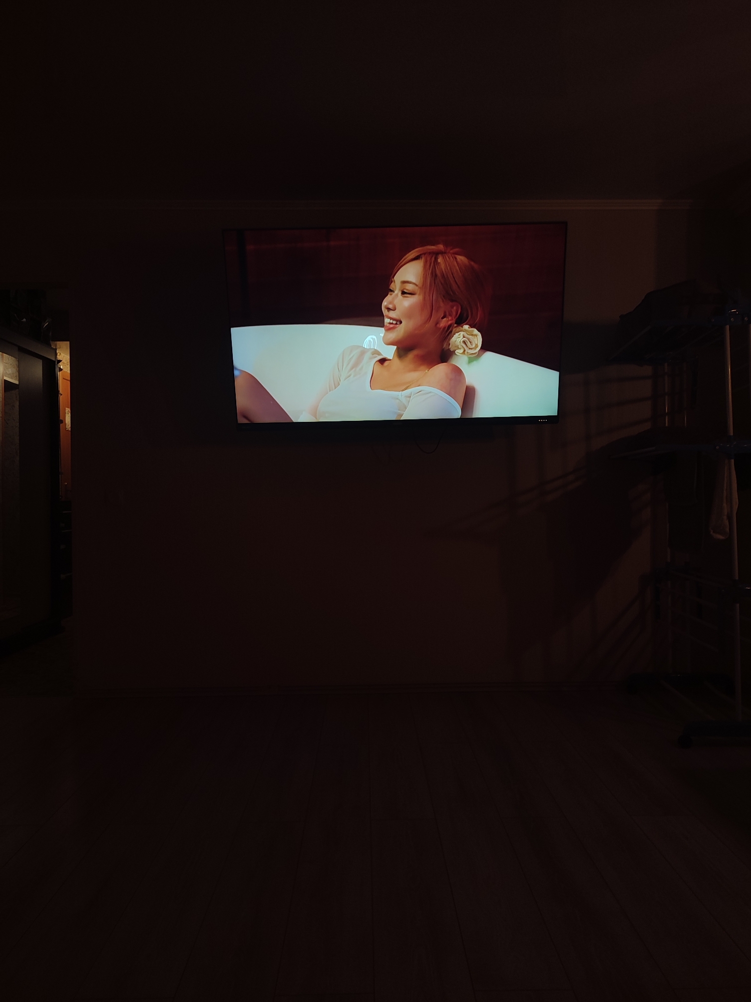 A woman with orange hair is shown on a television screen mounted on a wall. She is smiling and wearing a white top, with a white flower in her hair. The room is dimly lit, with wooden flooring visible. A clothes drying rack is present on the right side of the frame. The image appears to have been taken indoors in an apartment. It is not possible to determine the specific location in the city of Vsevolozhsk, Russia from the image alone.