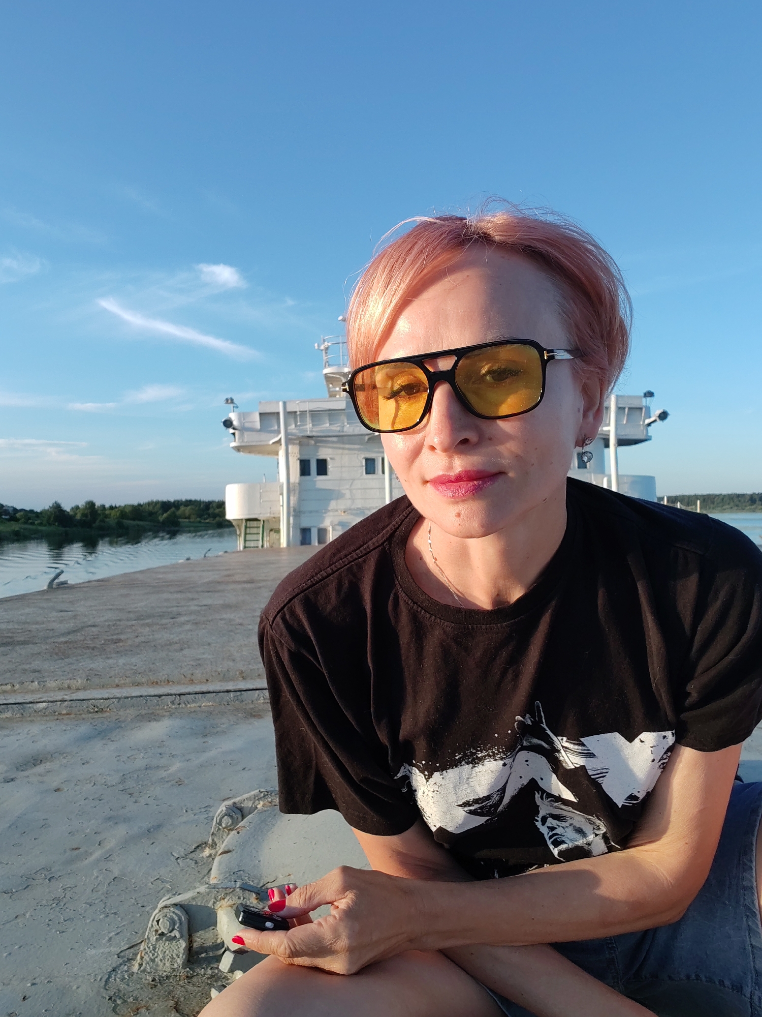 A woman with short, pink-hued hair and yellow-tinted sunglasses poses for a selfie outdoors. She is wearing a black t-shirt with a white graphic print and blue denim shorts. Her left hand holds a small black object with a red detail, her nails painted red. She is seated on a textured, metallic surface, possibly the deck of a boat or ship, with a body of water and a large white vessel in the background. The sky is bright blue with scattered wispy clouds, suggesting a sunny day. The light indicates it is either late afternoon or early morning. The scene appears to be a serene, outdoor setting, possibly on a river or canal, given the presence of the waterway and the docked ship.