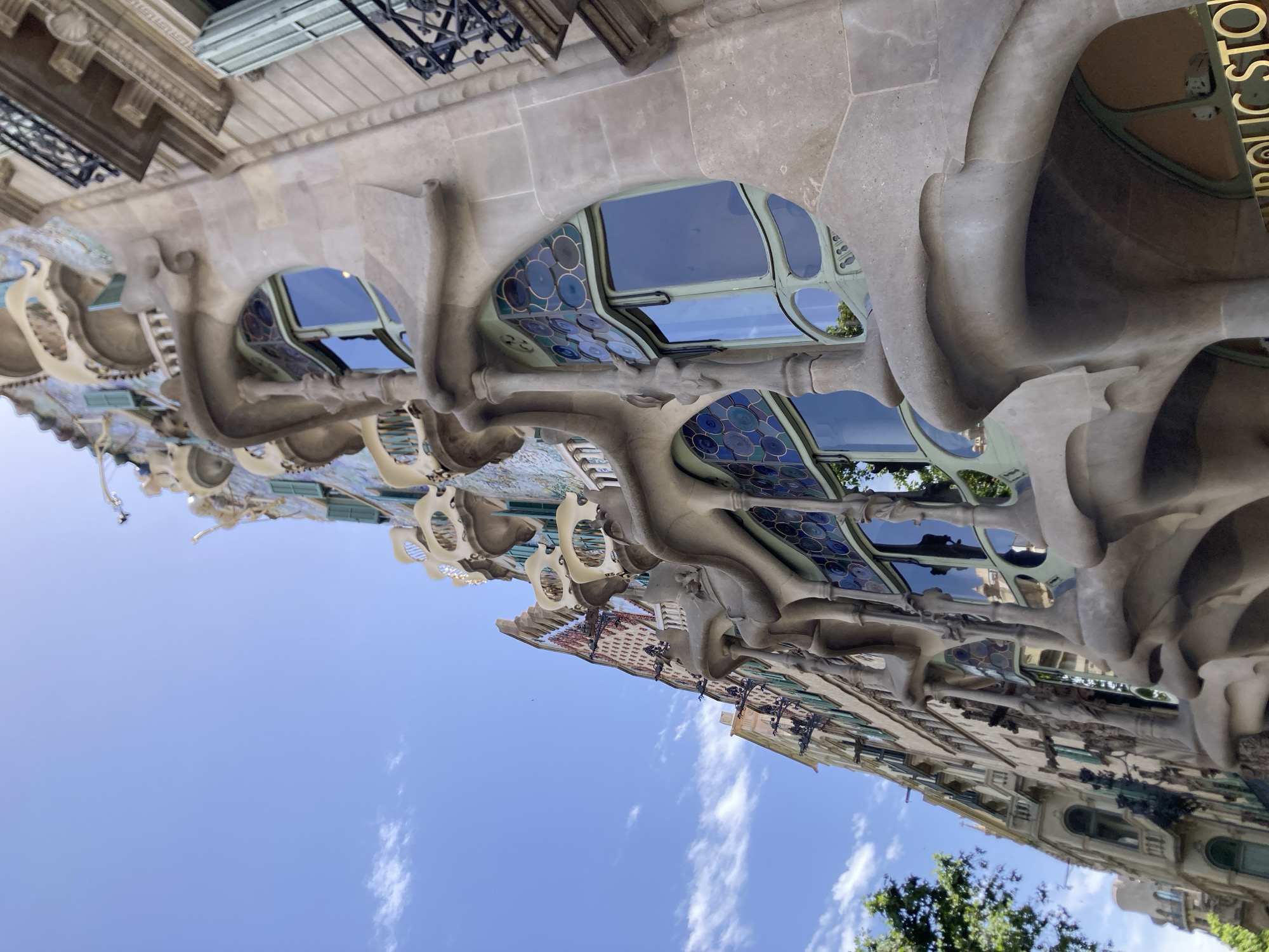 The image shows a close-up, low-angle view of the facade of Casa Batlló, a modernist building designed by Antoni Gaudí, located on Passeig de Gràcia in Barcelona, Spain. The building's distinctive organic, undulating facade is captured, featuring curved balconies that resemble bone-like structures and windows with colorful, circular stained glass. The sky above is blue with scattered white clouds. The sunlight highlights the textured stone and mosaic tile details of the building. Adjacent buildings with more traditional architecture and wrought-iron balconies are visible to the right and in the background, suggesting an urban streetscape. Trees are visible on the left side of the frame. No people are visible in the image. The overall impression is one of architectural artistry and vibrant design.