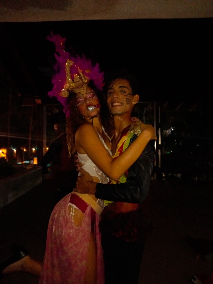 Two individuals in elaborate costumes are captured in an embrace against a dark, nocturnal background, indicative of an outdoor setting in the city of Playa El Agua, Venezuela. The female on the left wears a large fuchsia feathered and gold-sequined headpiece, with glittery eye makeup and a small white object held between her lips. She has on a white, possibly sequined, top and a pink, sheer skirt featuring a high slit revealing a red garment embellished with white beads. Her left foot is partially lifted, and she has her right arm around the male. The male on the right displays black facial markings on his right cheek and temple. He wears a dark, long-sleeved jacket over a multi-colored garment (red, yellow, green), dark pants, and a reddish-brown sash at his waist. He smiles directly at the camera, with both arms wrapped around the female. The background is dark and out of focus, with indistinct artificial lights visible, suggesting an evening event or performance.