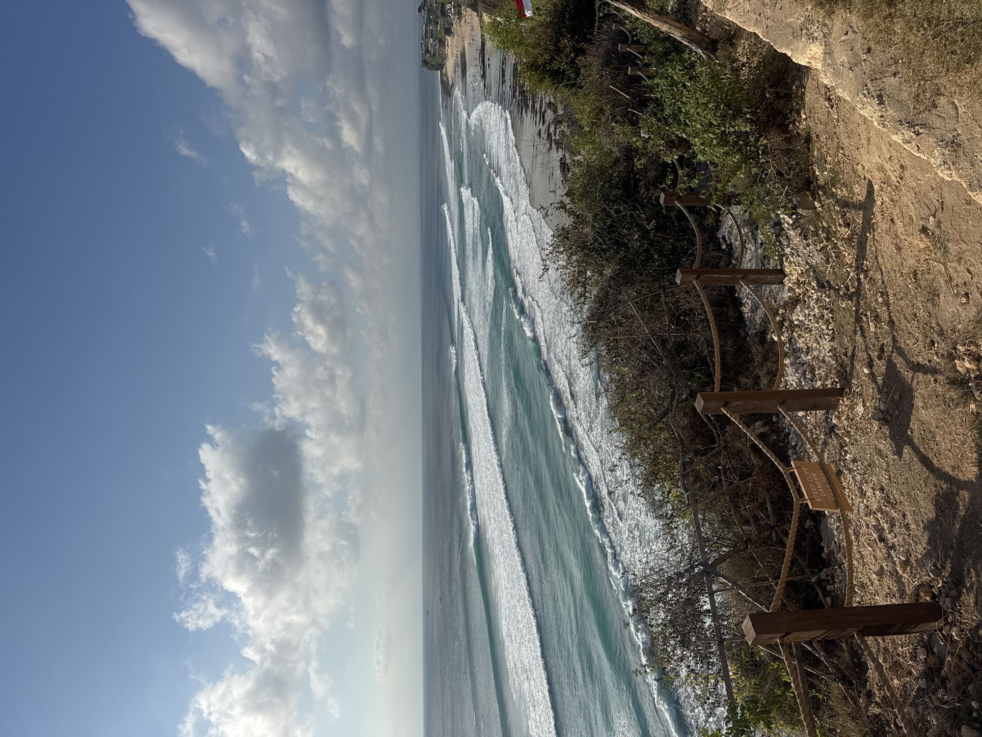 This is a medium-shot, eye-level, outdoor photograph of the beach at Uluwatu, located in Kuta Selatan, Bali, Indonesia. The sky is a bright blue with scattered, fluffy white clouds. The ocean is a vibrant turquoise, with multiple sets of waves rolling towards the shore. The waves appear to be breaking, forming white foam lines as they approach the beach. The beach itself is a mix of sand and rocky outcroppings. In the distance, there are buildings along the coastline, and numerous people are visible on the beach and in the water, some of whom may be surfing. In the foreground, a dirt path leads down towards the beach, bordered by a rustic wooden fence with rope railings. A small wooden sign is attached to the rope railing, with text that reads "STEEP CLIFF DANGER AWAS TEPI." Lush green vegetation and shrubs are growing along the cliff edge, obscuring parts of the view. The lighting suggests it is daytime, possibly late afternoon, with shadows cast by the vegetation and fence.