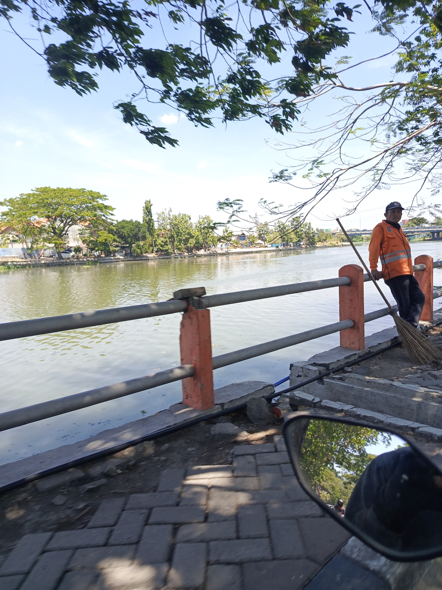 A street cleaner in an orange vest and black cap is standing next to a metal railing that borders a body of water. He is holding a broom and leaning on the railing. The scene appears to be a riverside promenade in Surabaya, Indonesia, with trees and buildings visible across the water. The sky is clear and blue. The foreground shows a paved walkway and a motorcycle's rearview mirror reflecting the scene.