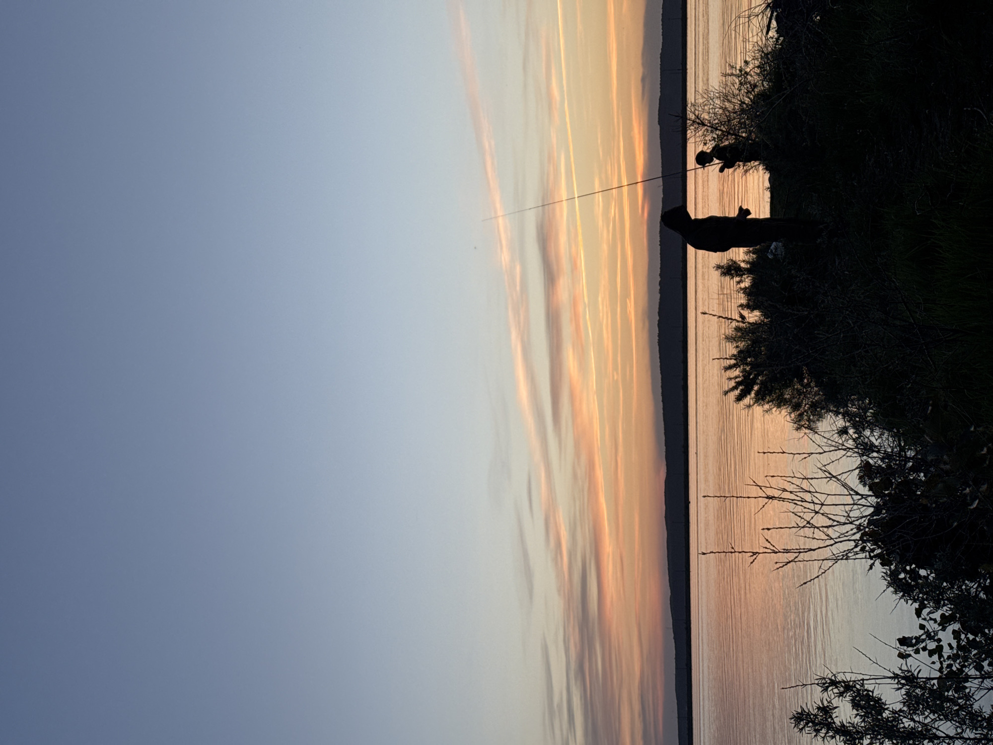 The image shows a twilight scene at a body of water in Chaikovsky, Russia. Two figures are silhouetted on the right side of the frame amidst vegetation. One appears to be an adult standing and holding a fishing rod, while the other seems to be a smaller person crouched nearby. The fishing rod extends across the water and into the sky. The sky is a gradient of blue to orange, reflecting in the water.