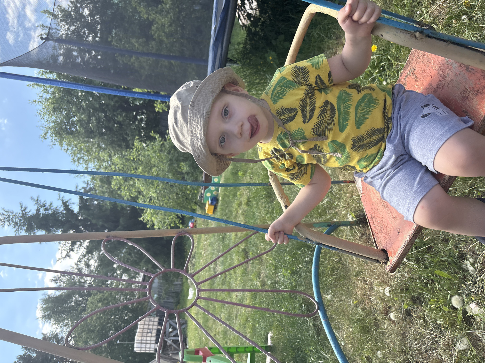 A young child wearing a tan bucket hat, a yellow shirt with a dark green fern pattern, and gray shorts with a zebra image on the leg sits on a swing. The swing has a rusty, red seat and is surrounded by a blue and light brown metal frame with a flower-shaped decoration. The swing set is positioned on grassy ground with dandelions. In the background, there is a trampoline with black netting, trees, and other playground equipment. The location is likely in Chaikovsky, Russia.
