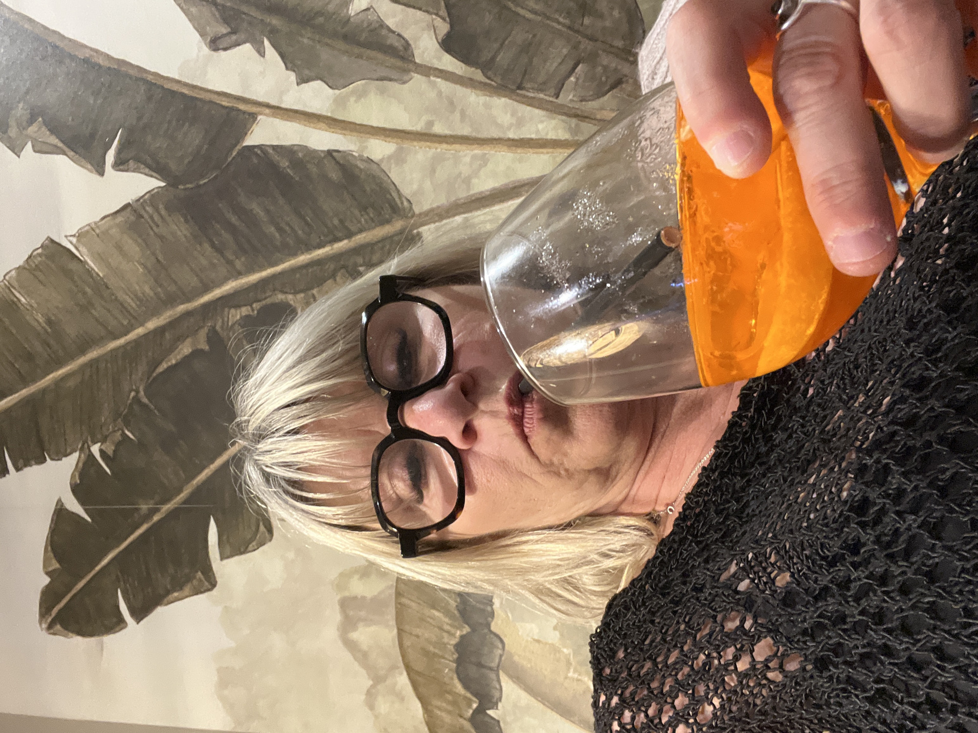 A woman is holding a glass of orange liquid up to her lips, preparing to take a sip. She has short blonde hair and is wearing black-rimmed glasses. She is wearing a black open-knit top and a ring on her finger. The background features a wall with a wallpaper pattern of large, stylized leaves. The location is not specified in the image; therefore, it is not possible to confirm this is in Grimaud, France.