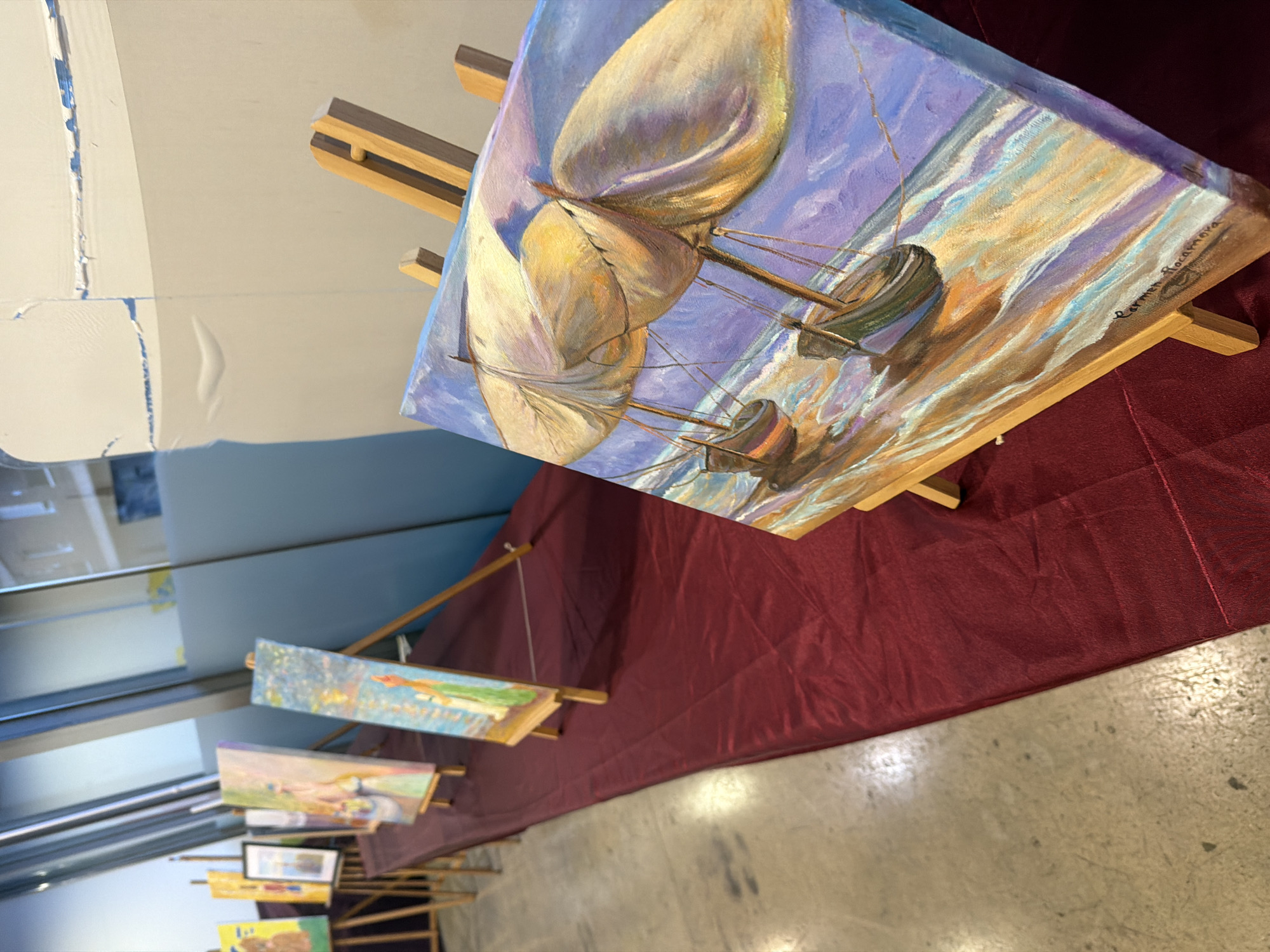 The image shows an art exhibition, likely indoors. Several paintings are displayed on easels along a long, red-clothed table. The closest painting in the foreground depicts sailboats with large sails on a body of water, rendered in vibrant blues, purples, and yellows, with a signature that appears to read "Carmen Rocamora." Other paintings further down the table are less clear but show varied subjects, including figures and cityscapes. The background suggests a modern building with large windows or glass partitions, and a concrete floor. The lighting appears to be artificial. There is no indication of the specific location in Elche/elx, Spain within the image itself.