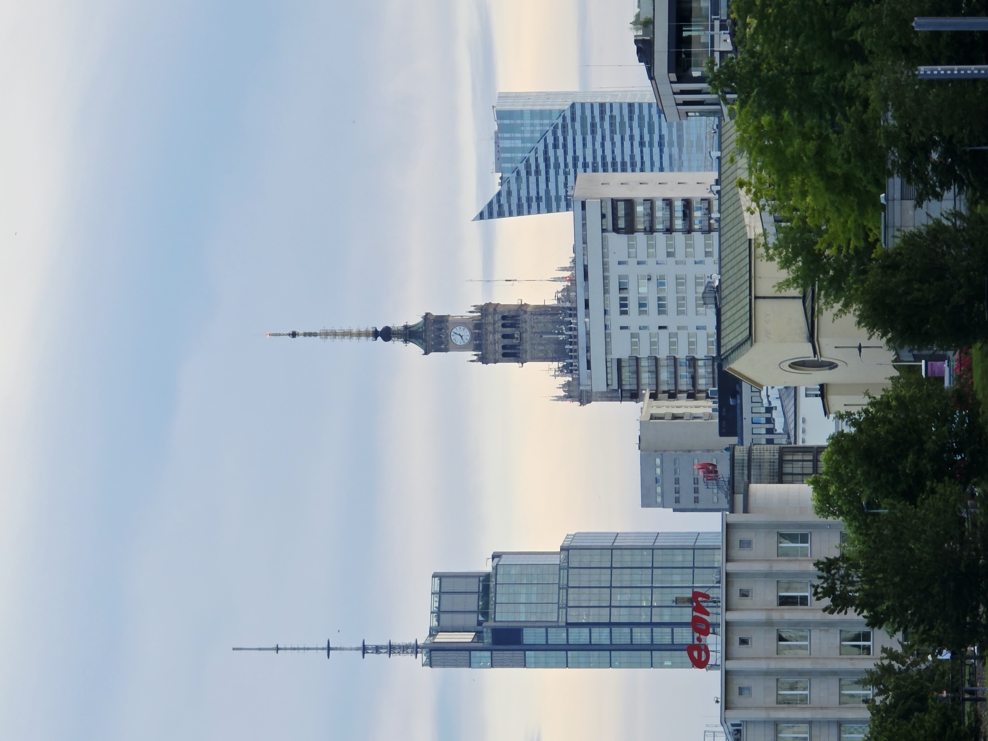 The image depicts a cityscape in Warsaw, Poland, featuring several prominent skyscrapers. In the foreground, trees partially obscure lower buildings. To the left, a tall, modern glass skyscraper with a distinctive antenna on top stands out. The word "e-on" is visible in red on the facade of a building in front of this skyscraper. Central to the image is the Palace of Culture and Science, a historic landmark with a clock tower. To the right of the Palace of Culture and Science, a contemporary glass building with a unique angled facade is visible. The sky above is overcast with scattered clouds. The overall scene is a blend of historical and modern architecture, characteristic of the Warsaw skyline. There are no visible people or discernible actions.