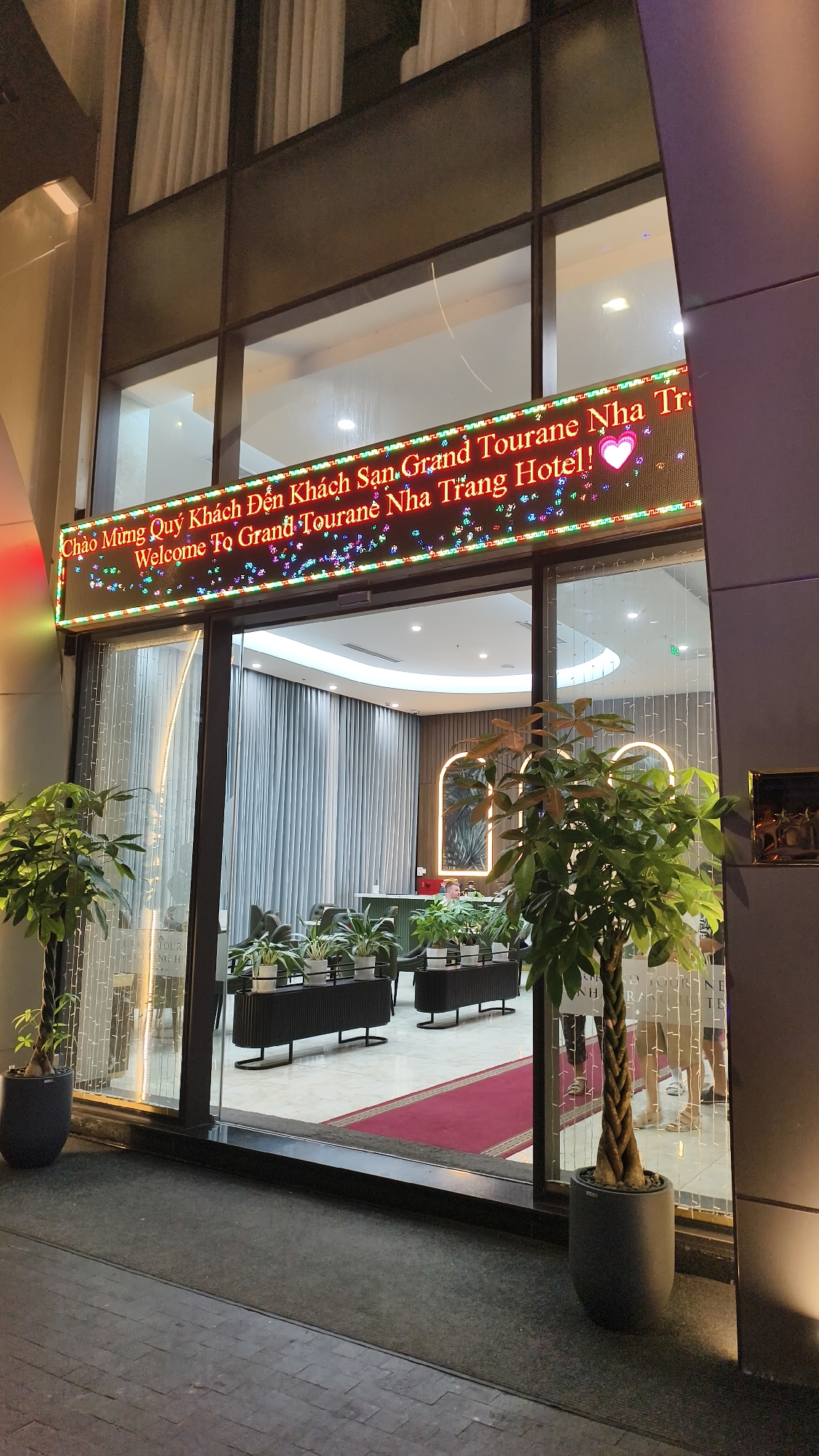 The image shows the exterior entrance of the Grand Tourane Nha Trang Hotel, located in Nha Trang, Vietnam. A brightly lit LED sign above the entrance displays a welcome message in Vietnamese and English: "Chào Mừng Quý Khách Đến Khách San, Grand Tourane Nha Trang Hotel! Welcome To Grand Tourane Nha Trang Hotel!". A pink heart symbol is also displayed on the sign. The entrance features large glass doors, with potted plants flanking the entryway. Inside, the hotel lobby is visible, with a reception desk, seating area, and decorative curtains. The flooring appears to be tiled, with a red carpet runner leading towards the interior. The scene is captured at night, indicated by the artificial lighting within and around the hotel.