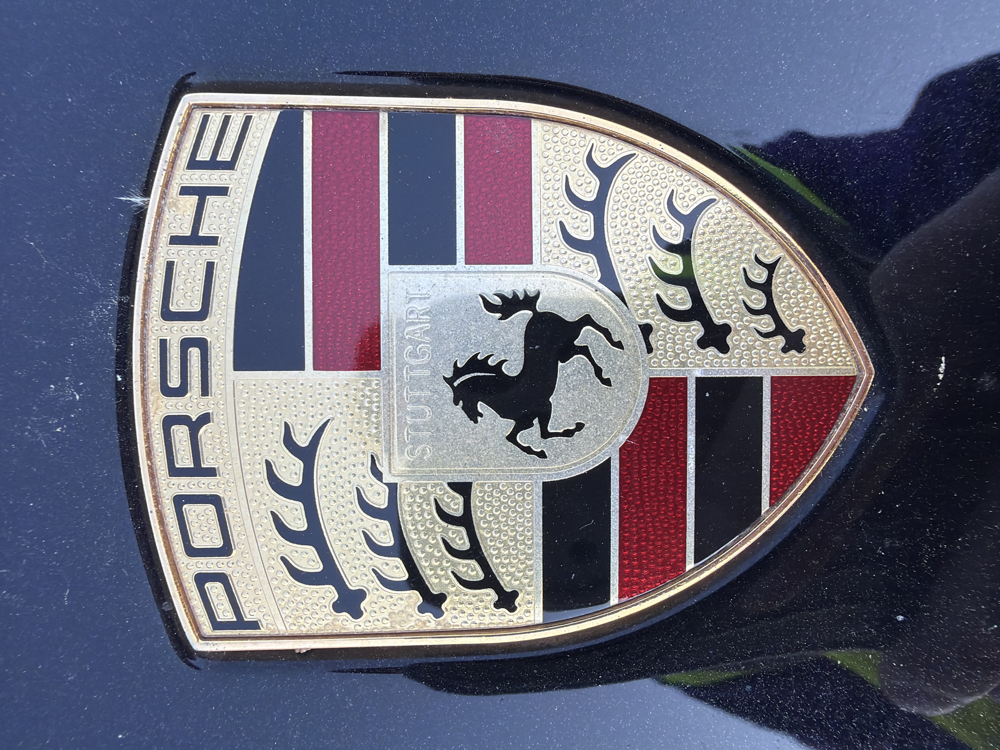 Close-up of a Porsche emblem affixed to a dark-colored vehicle. The emblem is shield-shaped with the word "PORSCHE" vertically aligned on the left side. The shield itself features a complex design with elements including a black horse rearing on its hind legs over the word "STUTTGART" and other heraldic symbols in gold, red, and black. The environment is the dark surface of the car, appearing slightly dirty. The location is not discernible from the image, and there is no information to indicate that the photo was taken in London.