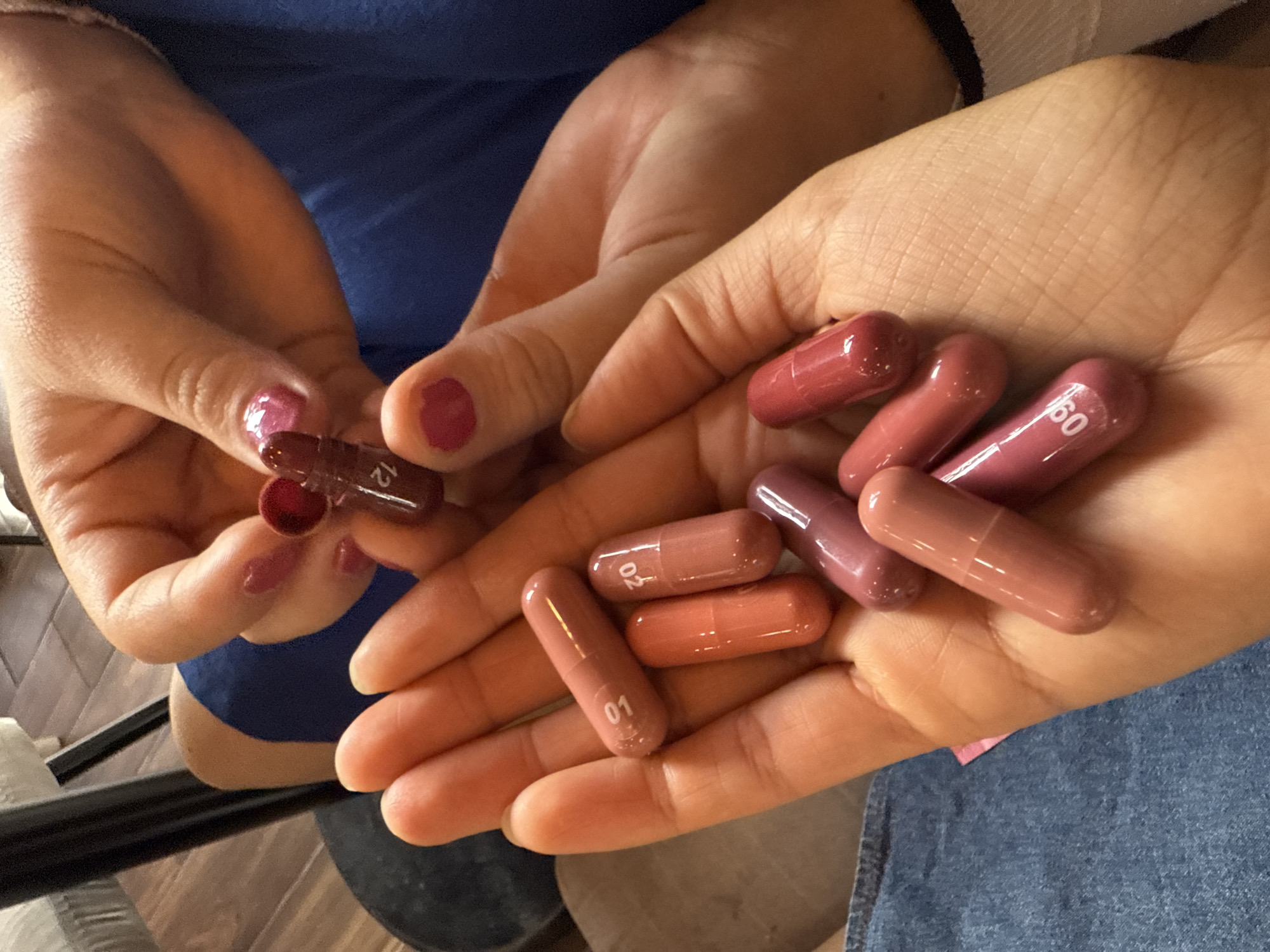 A person is holding a collection of reddish-brown capsules, some of which have numbers printed on them such as "01", "02", "12", and "60". The capsules are in the person's hands, with one hand holding two halves of an opened capsule. The person is wearing a blue shirt and sitting on a wooden chair. A snippet of light blue jeans can be seen in the bottom right of the image. The setting appears to be indoors. There is no evidence to place the location in Puebla, Mexico.