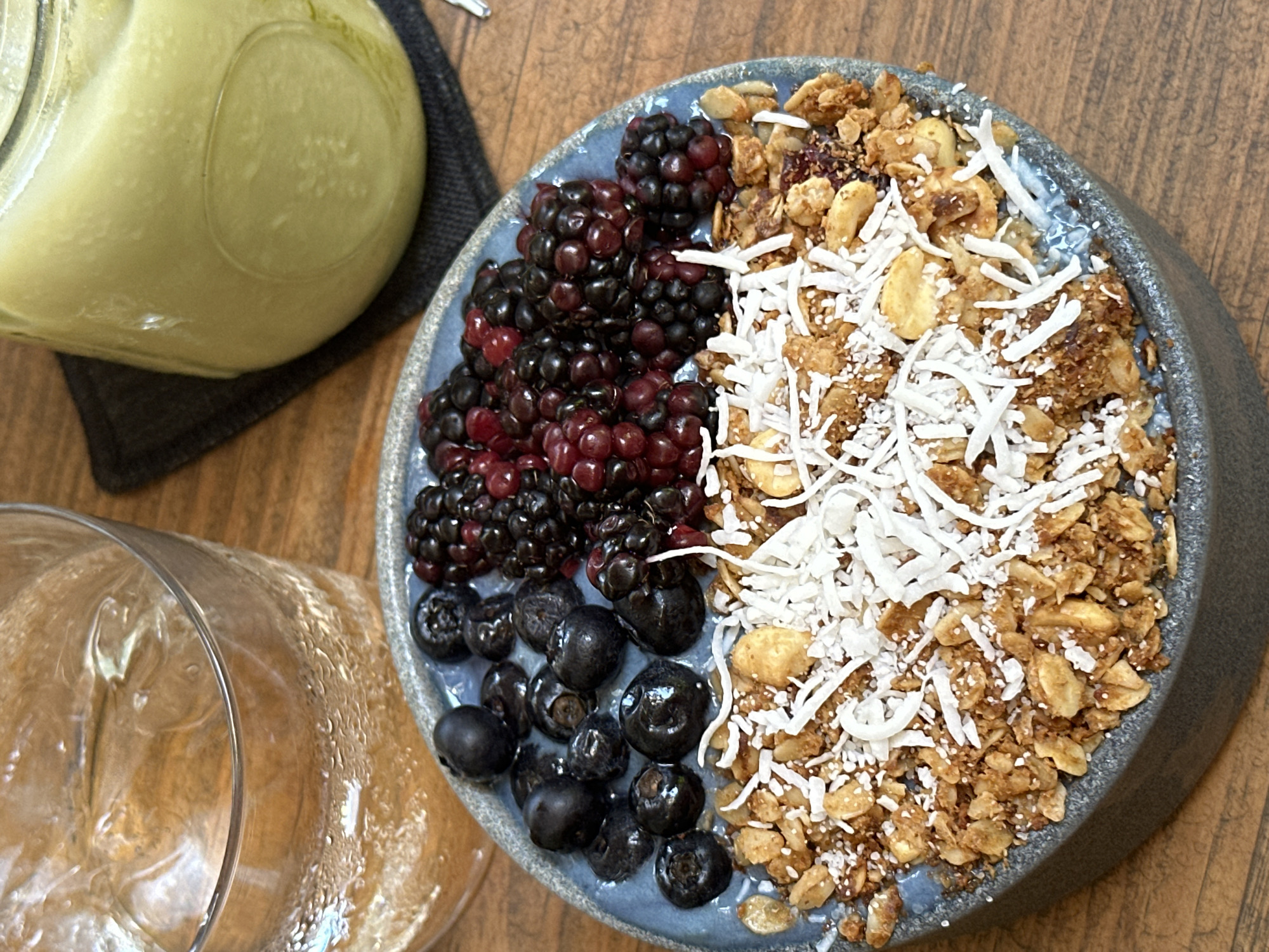 A close-up shot depicts a blue-gray bowl filled with what appears to be a smoothie bowl, featuring a combination of blackberries, blueberries, granola, and shredded coconut. To the left of the bowl, a jar containing a light yellow liquid and a glass are visible on a wooden surface. The specific location within Puerto Escondido, Mexico, cannot be determined from this image.
