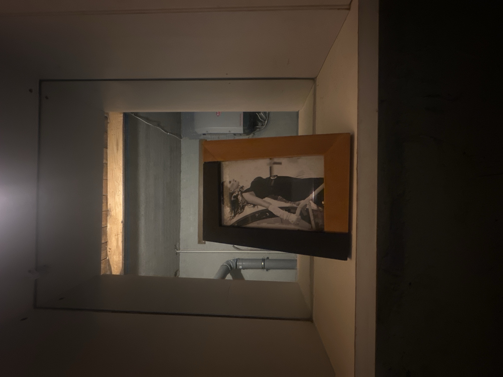 The photograph captures a framed black and white image of a woman in a dress and gloves, set against a dark backdrop. This frame, with its wooden and black elements, rests on a light-colored shelf. The shelf appears to be within a niche or recessed area, flanked by plain walls. Beyond the shelf and the framed picture, a view into another space is visible. This space reveals a concrete ceiling with visible wooden beams, exposed pipes, and a gray electrical box. The overall scene suggests an indoor setting, possibly a basement or utility area, located in Lapės, Lithuania.