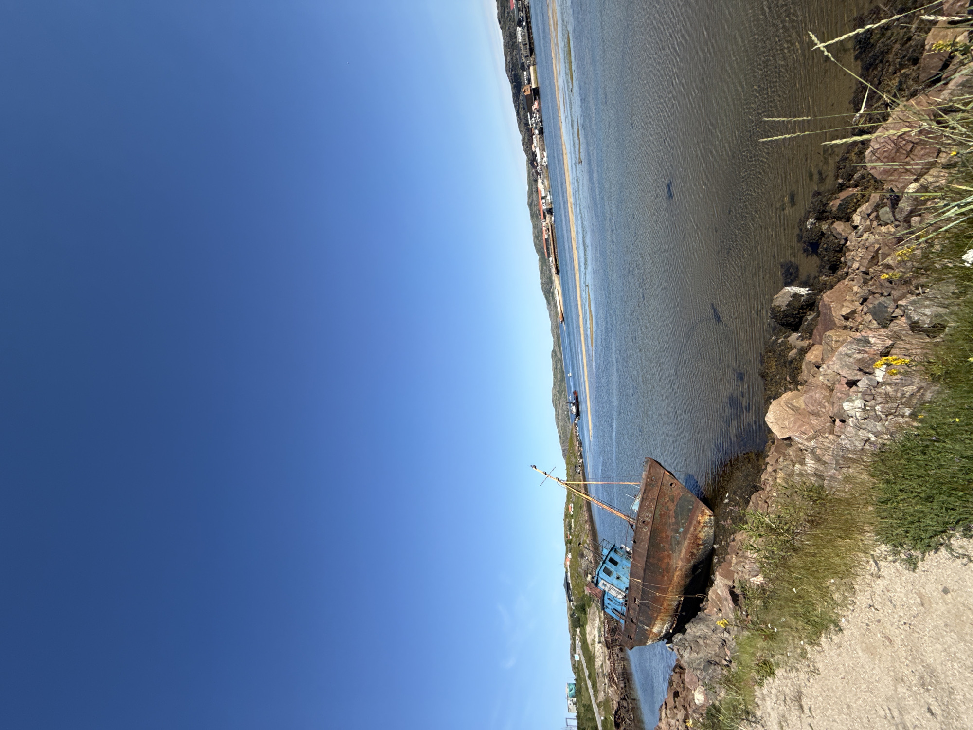 A rusty, abandoned fishing trawler with a blue cabin rests on its side on a rocky shore. The boat is tilted, with its stern submerged in the shallow, clear water. The sky is a bright, cloudless blue. In the background, across a body of water, is a settlement with buildings and more boats. The shore in the foreground is covered in stones and sparse vegetation. This scene is located in Teriberka, Russia, a coastal village on the Barents Sea, known for its dramatic landscapes and shipwrecks.
