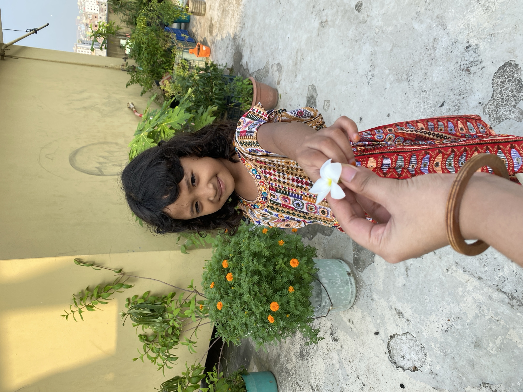 The image captures a moment on a rooftop in Dhaka, Bangladesh. A young girl with dark hair and a colorful dress is smiling as she receives a small white flower from a hand (presumably an adult) wearing a wooden bracelet. The girl is near a pot of marigolds. In the background, there are other potted plants, a yellow wall, and a cityscape. The ground is concrete.