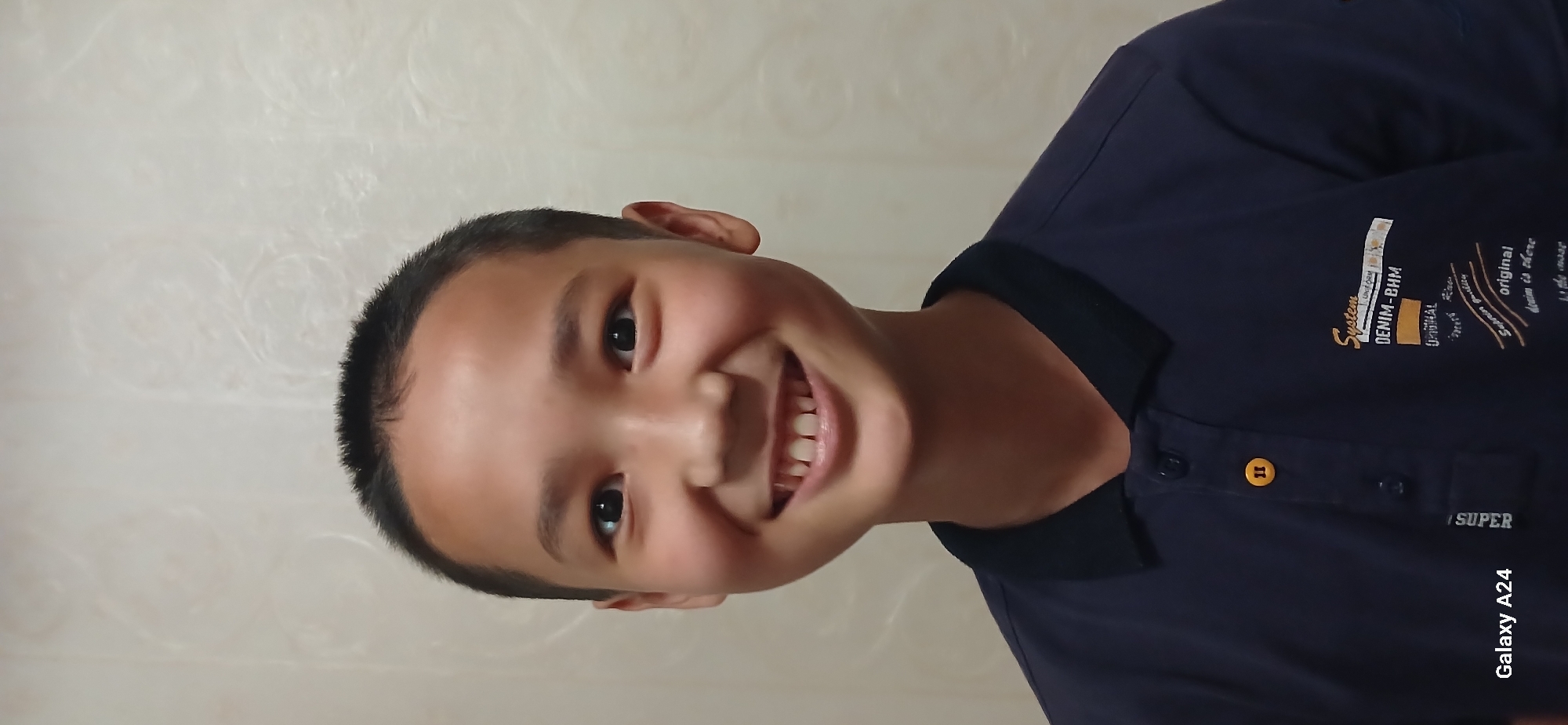 The image shows a close-up portrait of a young East Asian boy with a wide smile, revealing his teeth. He has short, dark hair and is wearing a dark blue polo shirt with a black collar and two buttons. A yellow button is visible on the placket, and a yellow symbol is also present on the shirt. Text is visible on the chest of the shirt, including "System", "DENIM-BHM", and "original". The background is a textured cream-colored wall. The bottom left corner of the image displays "Galaxy A24". The image appears to be taken indoors, with neutral lighting. There are no indications of the location being specifically in Bishkek City, Kyrgyzstan, or any other specific city.