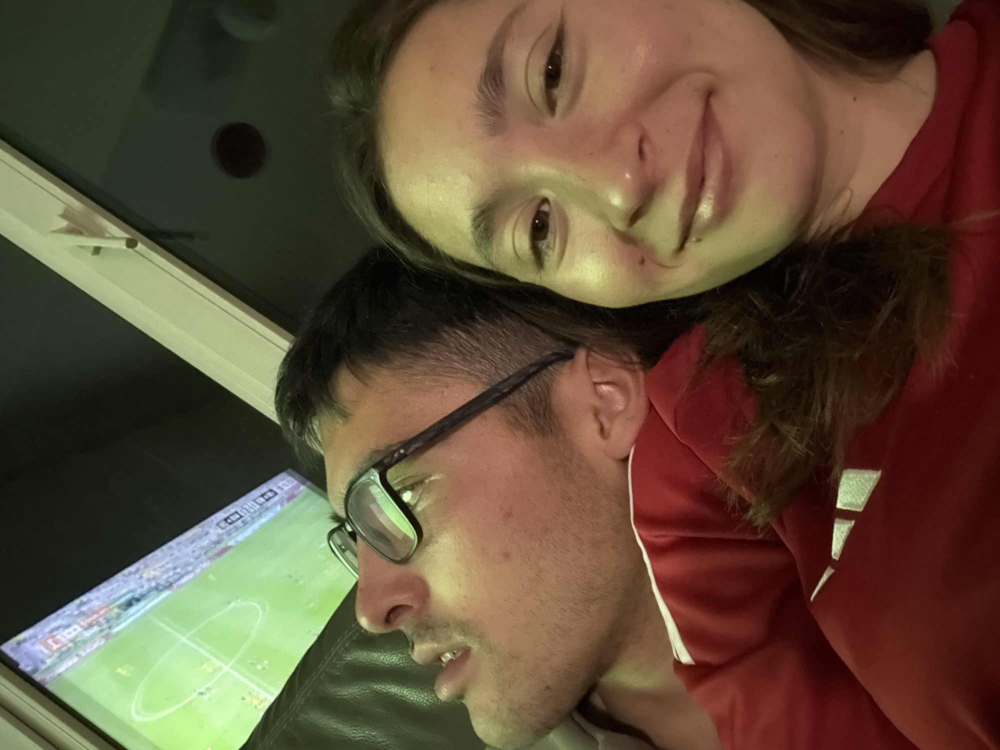 A couple is watching a soccer match on a television screen in what appears to be a living room. The man, wearing glasses and a red shirt, is on the left side of the frame, looking up at the television with his mouth slightly open. The woman is on the right side of the frame, smiling and looking directly at the camera. The television screen displays a soccer field with players. The room is dimly lit, with the light from the television casting a greenish hue on the scene. The image does not contain any information about the city of Sofia, Bulgaria.