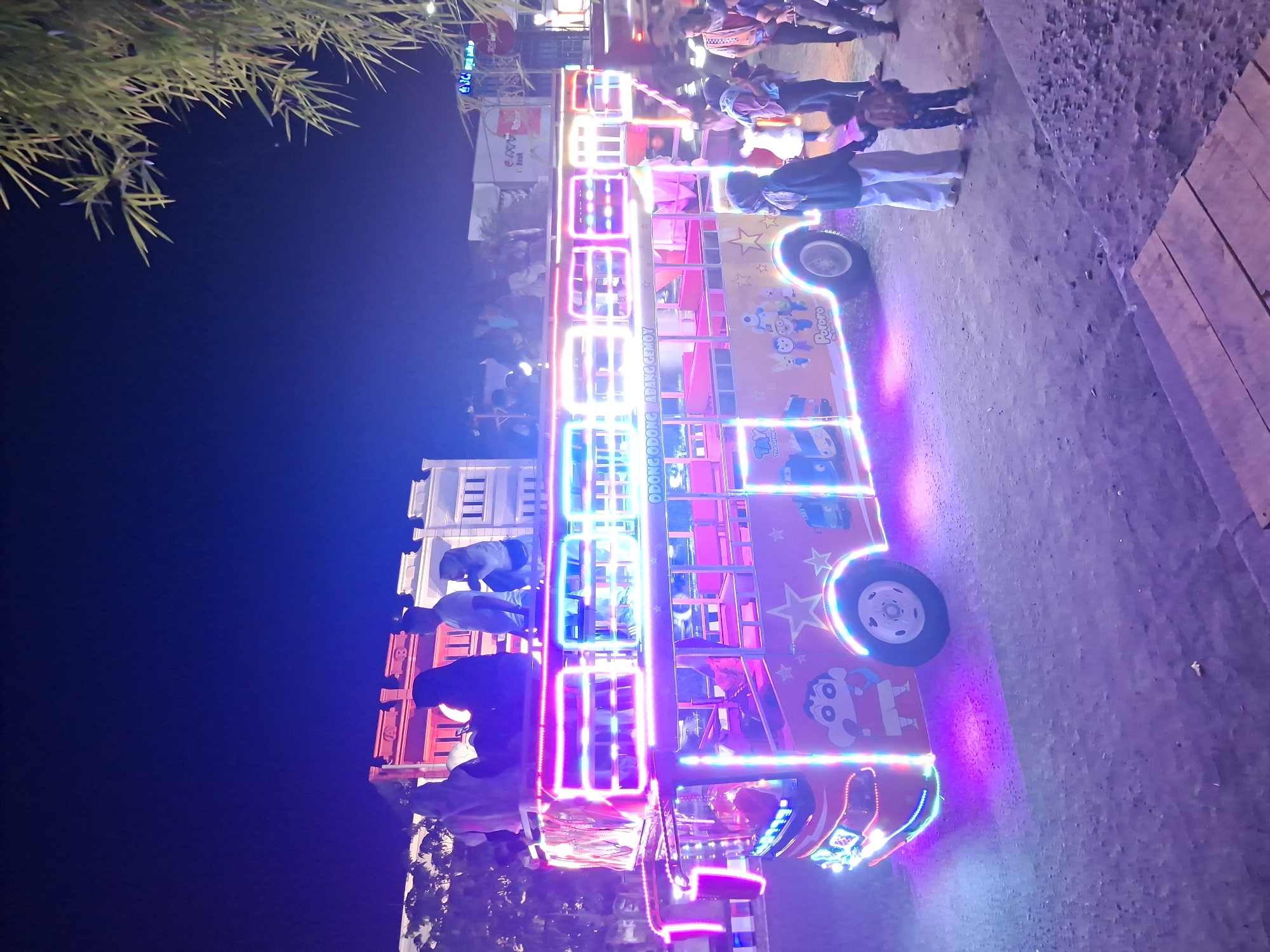 The image captures a vibrant night scene in Lhokseumawe, Indonesia, centered around a brightly illuminated double-decker bus. The bus, likely a modified vehicle for entertainment or a ride, is extensively decorated with colorful LED lights in pink, blue, white, and purple, creating a festive glow. Cartoon characters such as Pororo the Little Penguin, Tayo the Little Bus, and Crayon Shin-chan are painted on its sides, along with stars.
Several people, including adults and children, are present around and on the bus. Some individuals are visible on the upper deck, while others stand on the ground, suggesting they are either participants or spectators of this lively event. The ground surface appears to be dirt or gravel. In the background, multi-story buildings with illuminated windows are visible under a dark night sky, confirming the time of day. Lush, bamboo-like foliage is present in the upper left of the frame.
Visible text on the bus includes "ODONG ODONG ABANG GEMOY" prominently displayed on the lower deck. Other readable text includes "Pororo," "Tayo," and "Kiddy Bus." The overall atmosphere is one of a cheerful evening gathering or a fun fair, characterized by the bright lights and animated decorations of the bus.