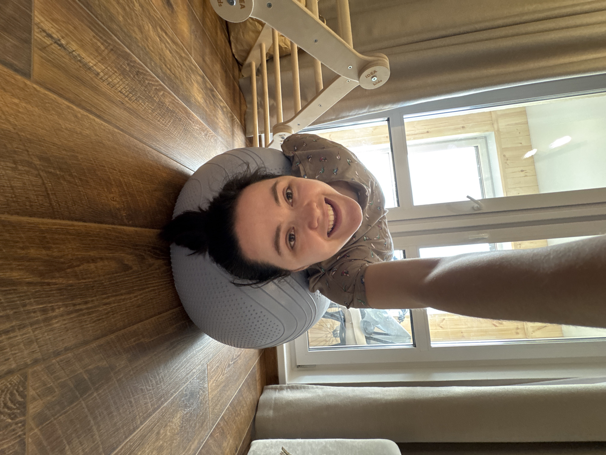 A woman with dark hair tied back is depicted in a rotated, low-angle selfie, lying on her back with her head resting on a large, textured light grey exercise ball. She wears a patterned long-sleeved top and smiles broadly at the camera, conveying happiness and ease.
The setting appears to be an indoor room in Izhevsk, Russia, characterized by a warm, medium-brown wooden floor with visible grain running diagonally across the left side of the frame. Bright daylight streams in through a prominent white-framed window on the right. Through the window, a wooden fence or structure is visible outside. Beige curtains frame the window area.
Above the woman, positioned towards the ceiling, is a light-colored wooden play structure, possibly a climbing frame or Pikler triangle, featuring rounded joints and rungs. The brand name "IDEA kids" is clearly visible on one of the circular wooden joint pieces of this structure. A rolled-up, light-colored blanket or cushion is also visible on the floor near the window. The overall impression is a bright, comfortable home environment during the daytime.
