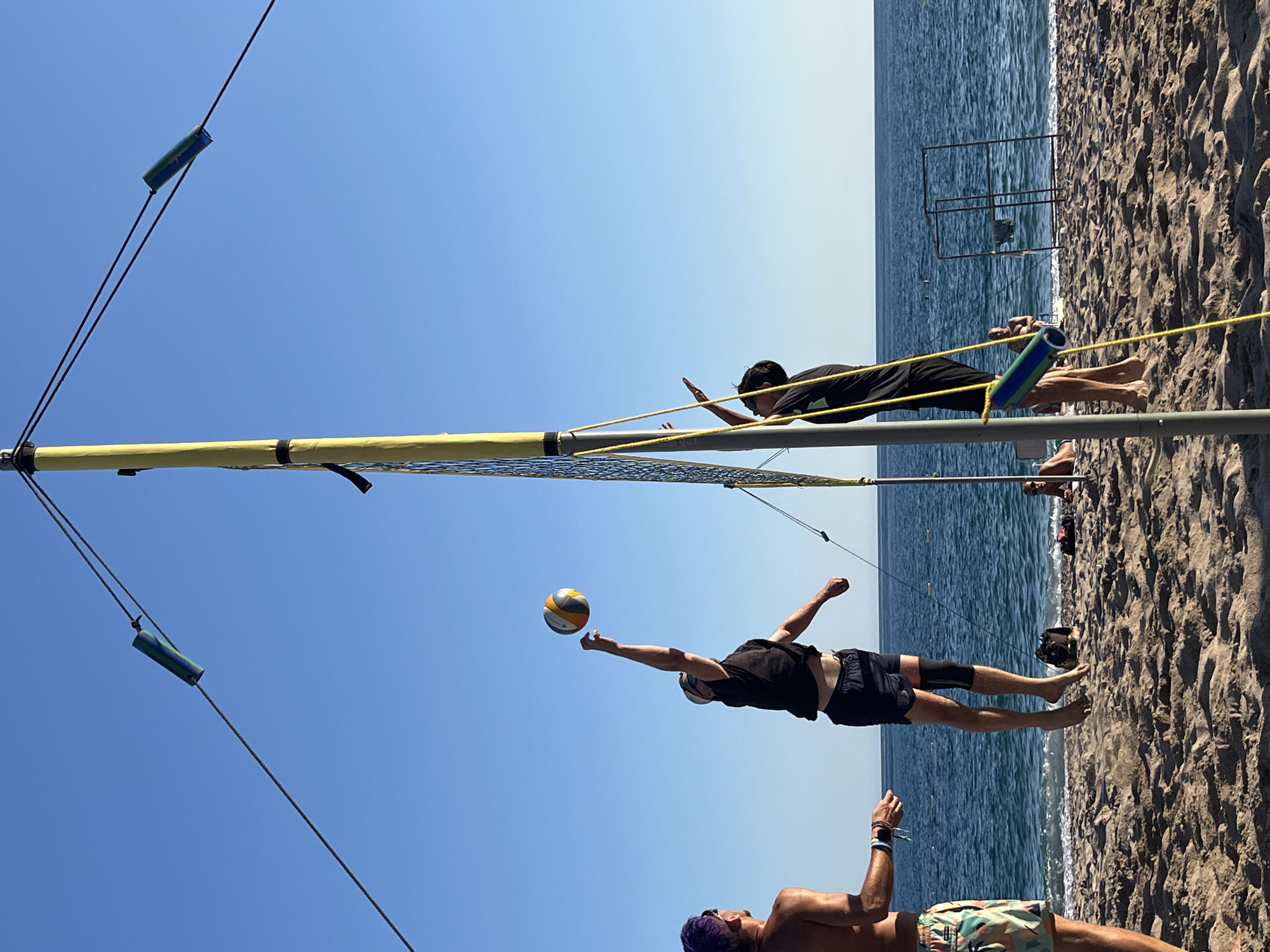 The image depicts a beach volleyball game taking place on a sandy beach in Zankovići, Montenegro. The scene is set against a clear blue sky and the calm blue sea. In the foreground, three men are actively engaged in the game. One man, wearing a black t-shirt and dark shorts with knee pads, is in mid-air, jumping to hit a volleyball that is positioned above his outstretched right hand. Another man, in the foreground to the left, wearing colorful patterned shorts and a purple-dyed hairstyle, is observing the play. A third man, wearing a black t-shirt and dark shorts, stands near the volleyball net, seemingly ready to receive a hit. The volleyball net is taut, supported by a yellow pole. Several other people are visible in the background, relaxing on the beach or near the water. The sand shows signs of activity, with footprints and disturbed areas. The overall environment is consistent with a coastal recreational area.