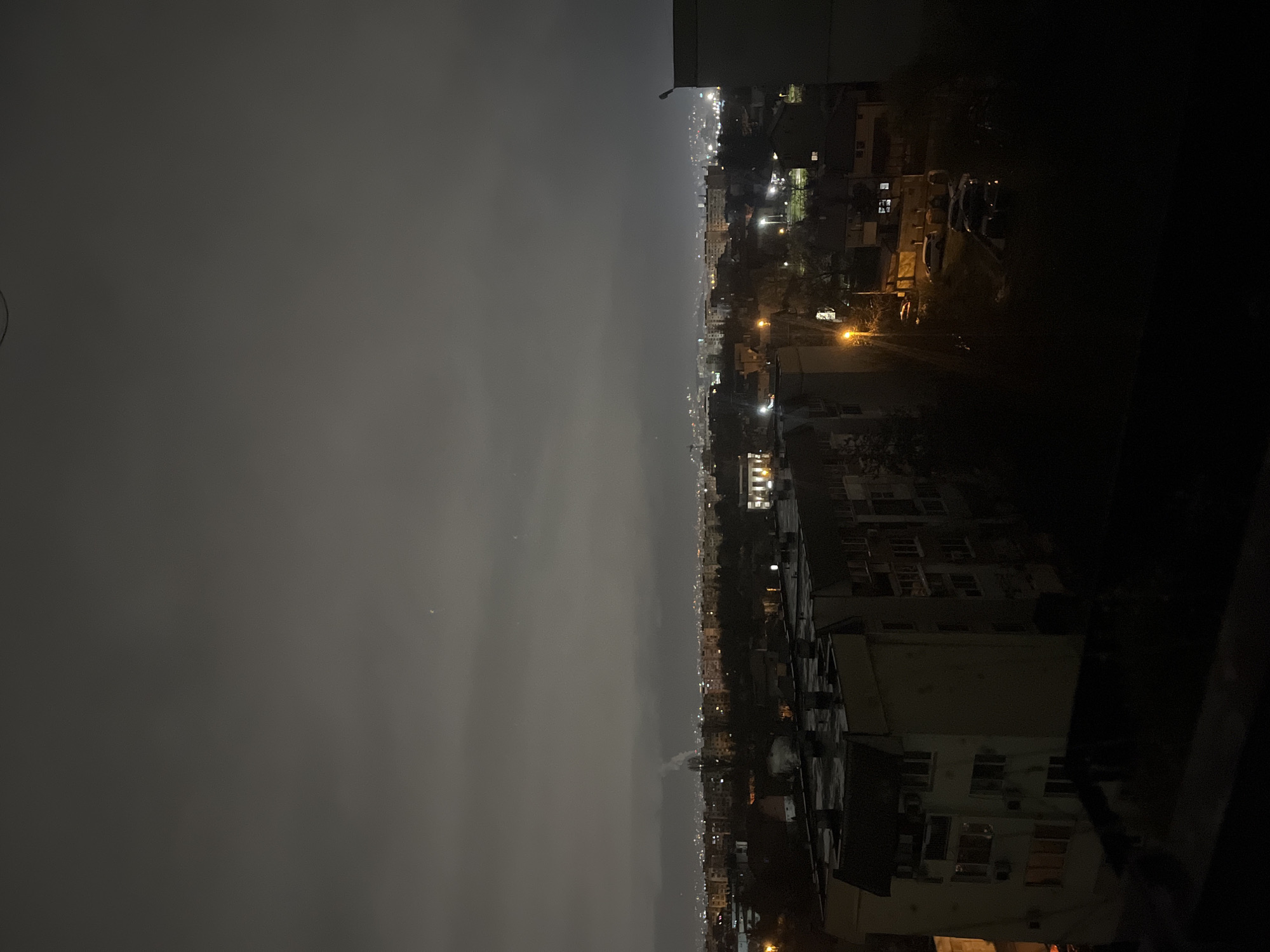 The image depicts a nighttime view from a high vantage point, likely a building, in Sofia, Bulgaria. The scene is split between a dark sky, appearing overcast or smoggy, on the left, and the city illuminated by streetlights and building lights on the right. The buildings closest to the viewer are dimly visible in the foreground. The lights of the city stretch into the distance, indicating a substantial urban area. There is smoke emitting from a chimney that is in the center of the image. There is no evidence of human presence, actions, or interactions directly visible within the frame.