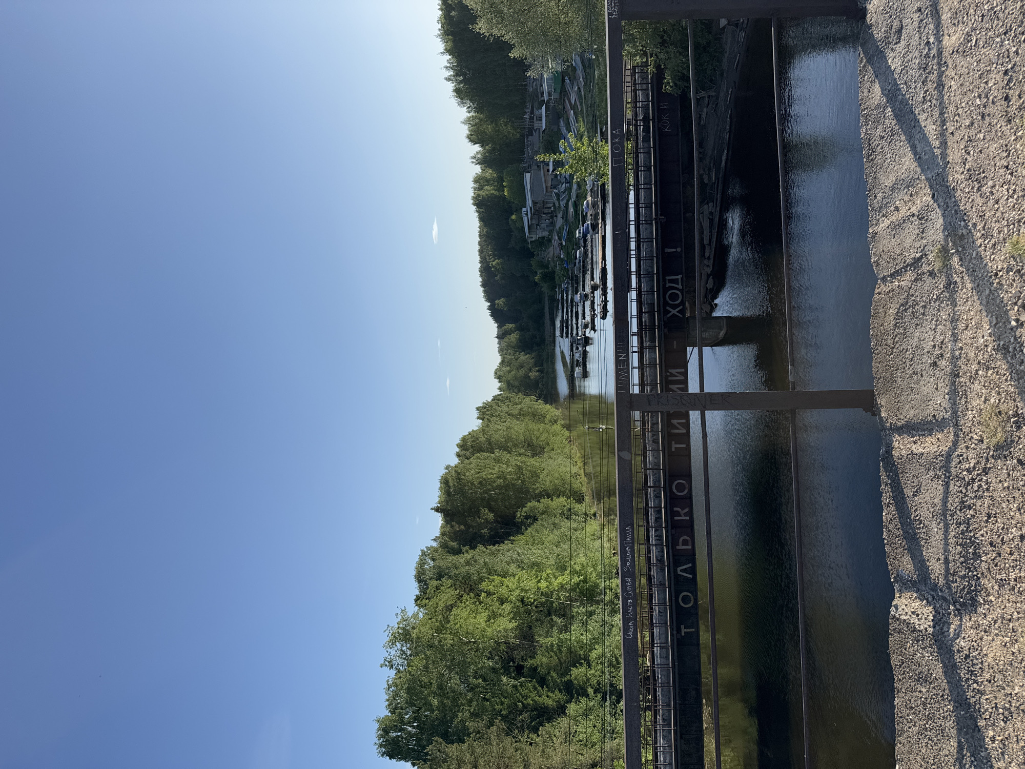 A daytime shot shows a view from a bridge in Chaikovsky, Russia. The bridge, built from steel and gravel, spans a river, lined on both sides with green trees. Across the river, a cluster of buildings is visible. The sky is blue. Russian text appears on the side of the bridge.