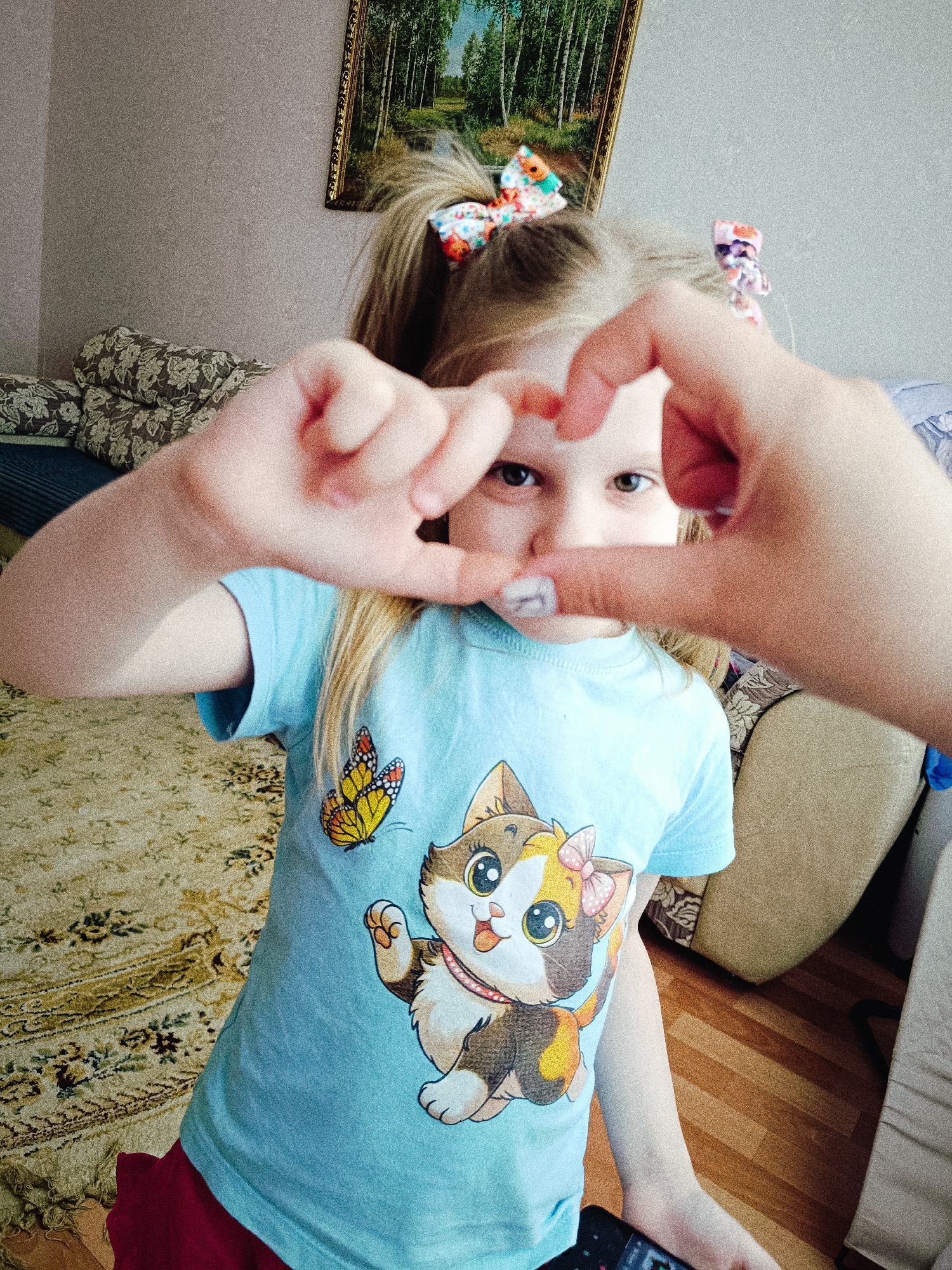 A young girl stands indoors, making a heart shape with her hands, and looking through the opening. She has blonde hair with two colorful bows. She is wearing a light blue T-shirt with a cartoon cat and butterfly on it, and red shorts. The background includes a patterned couch, a painting of a forest, light beige walls, and wooden flooring. The location is likely a residential setting in Tobolsk, Russia.