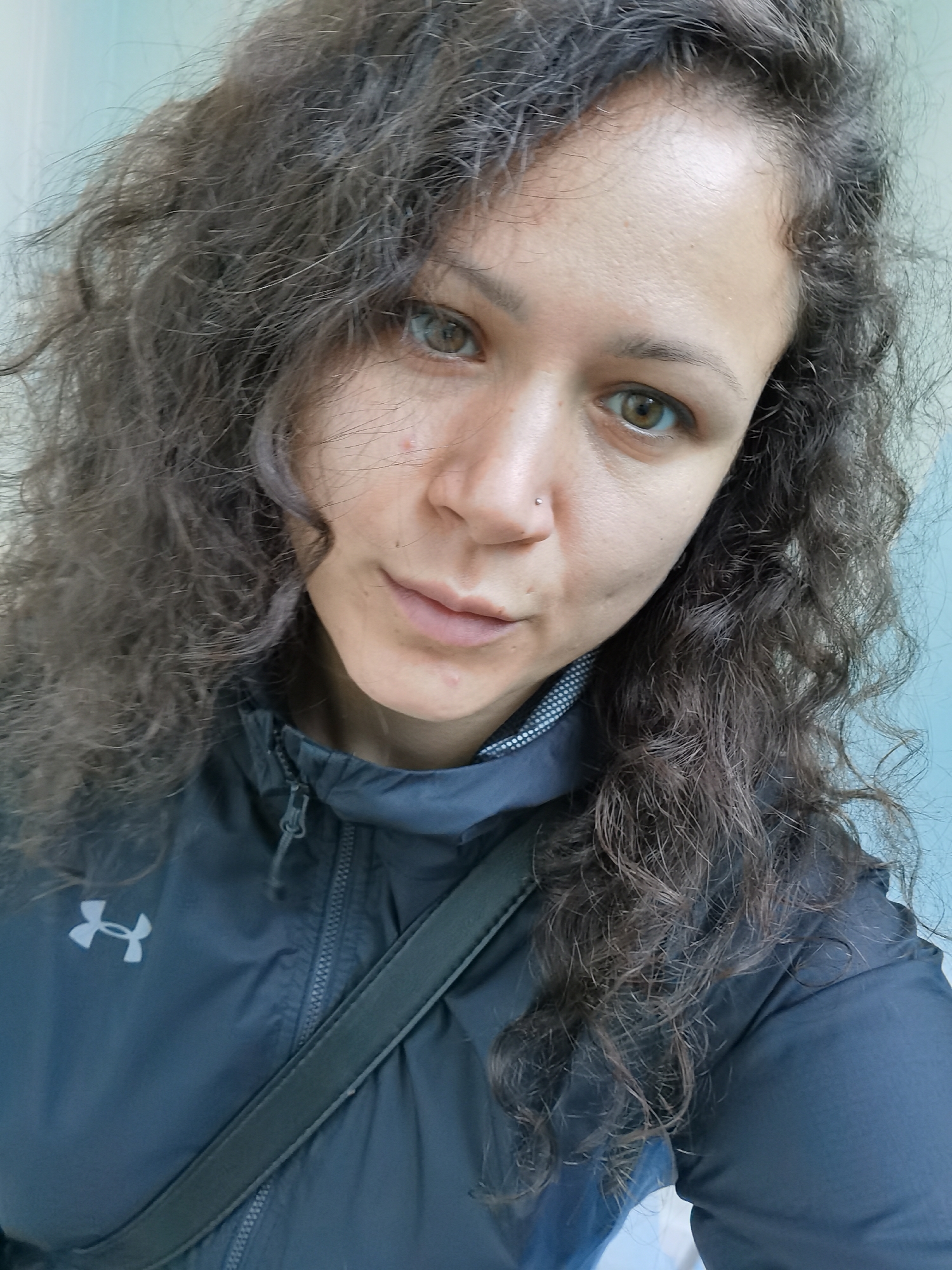 The image portrays a female individual located in the city of Chaikovsky, Russia, captured in a head-and-shoulders frame. The subject is an adult woman with long, dark brown, curly hair that appears slightly frizzy. She possesses light brown or hazel eyes and directly faces the viewer. A small, shiny, possibly silver or clear gemstone, stud piercing is visible on her left nostril. Minor skin imperfections, including small red marks, are present on her forehead and chin. She is dressed in a dark blue or black zip-up jacket. The jacket features a prominent white Under Armour logo on the left chest. The inner collar of the jacket exhibits a subtle gray and white patterned texture. A dark, flat strap, consistent with a bag strap, traverses diagonally across her upper torso. The background is softly blurred, displaying indistinct light blue and lighter, possibly off-white, surfaces.