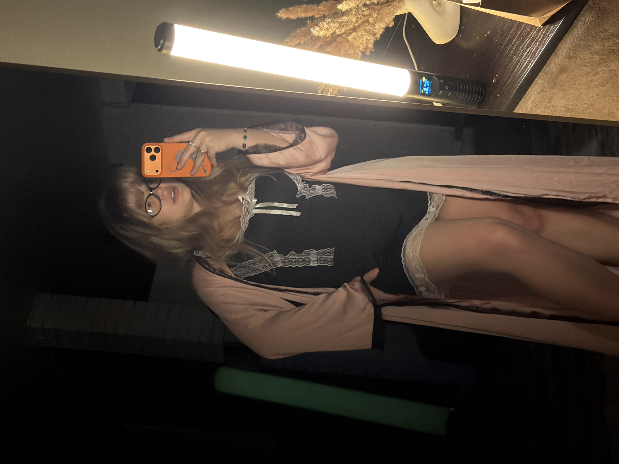 The image is a selfie taken by a person in a black and white lace-trimmed bodysuit and a pink robe, visible in a mirror. The person has blonde hair and is wearing glasses. They are holding an orange smartphone with multiple camera lenses. A light source is positioned in the upper portion of the frame, suggesting indoor lighting. A bedside table with decorative elements like dried plants and a lamp is partially visible on the upper right. The location is possibly inside a bedroom or dressing room in Большая Шильна, Russia. A green object is observed in the lower part of the image.