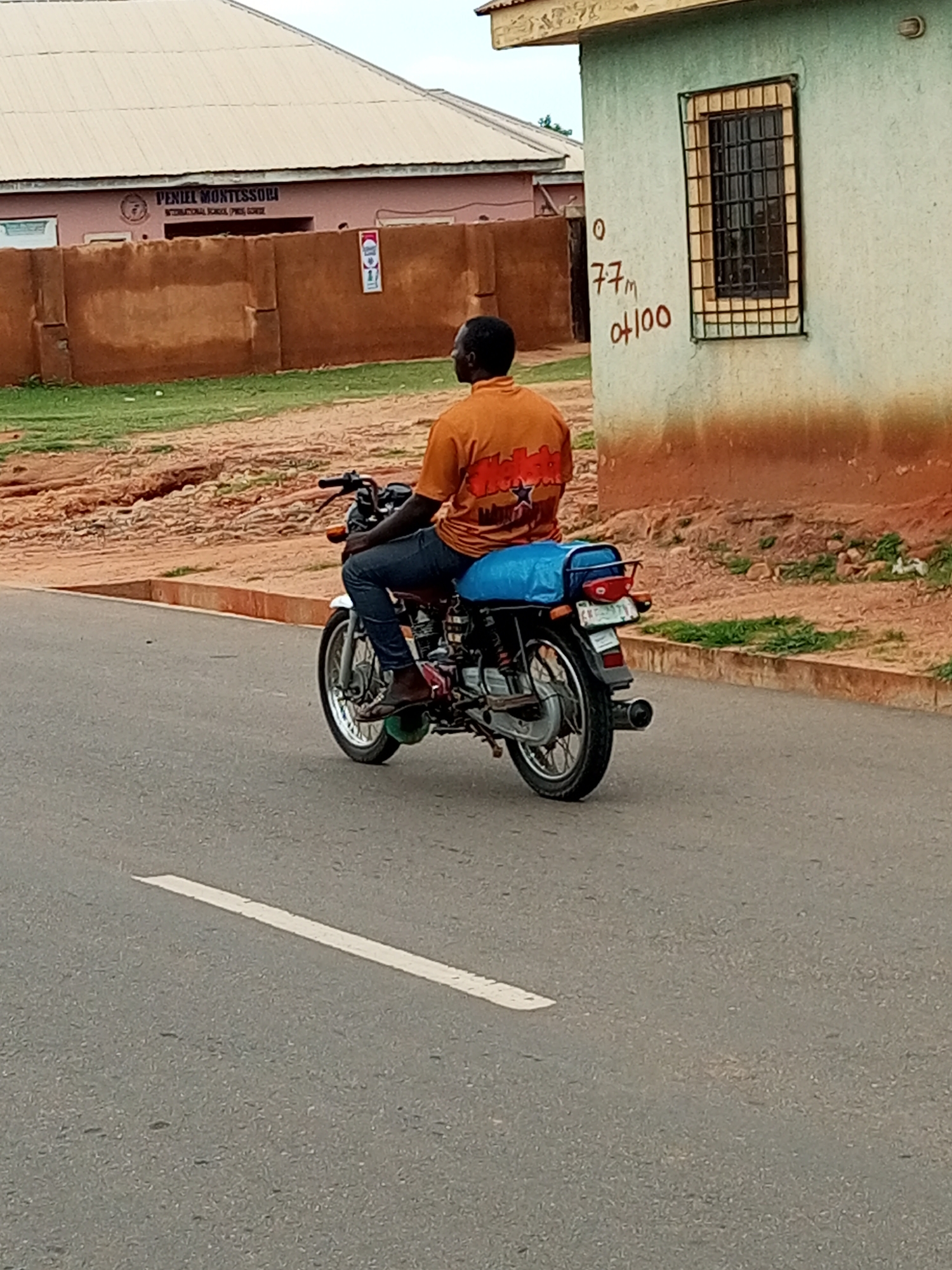 A person wearing an orange t-shirt and blue jeans rides a motorcycle on an asphalt road. The motorcycle has a blue tarp covering the back seat and a license plate. In the background, there is a building with a green facade and a barred window. To the left of that building, there is a pink building with the text "PENIEL MONTESSORI INTERNATIONAL SCHOOL" visible. The ground is covered in dirt and grass. The image appears to be taken in a developing country given the architecture and road conditions.