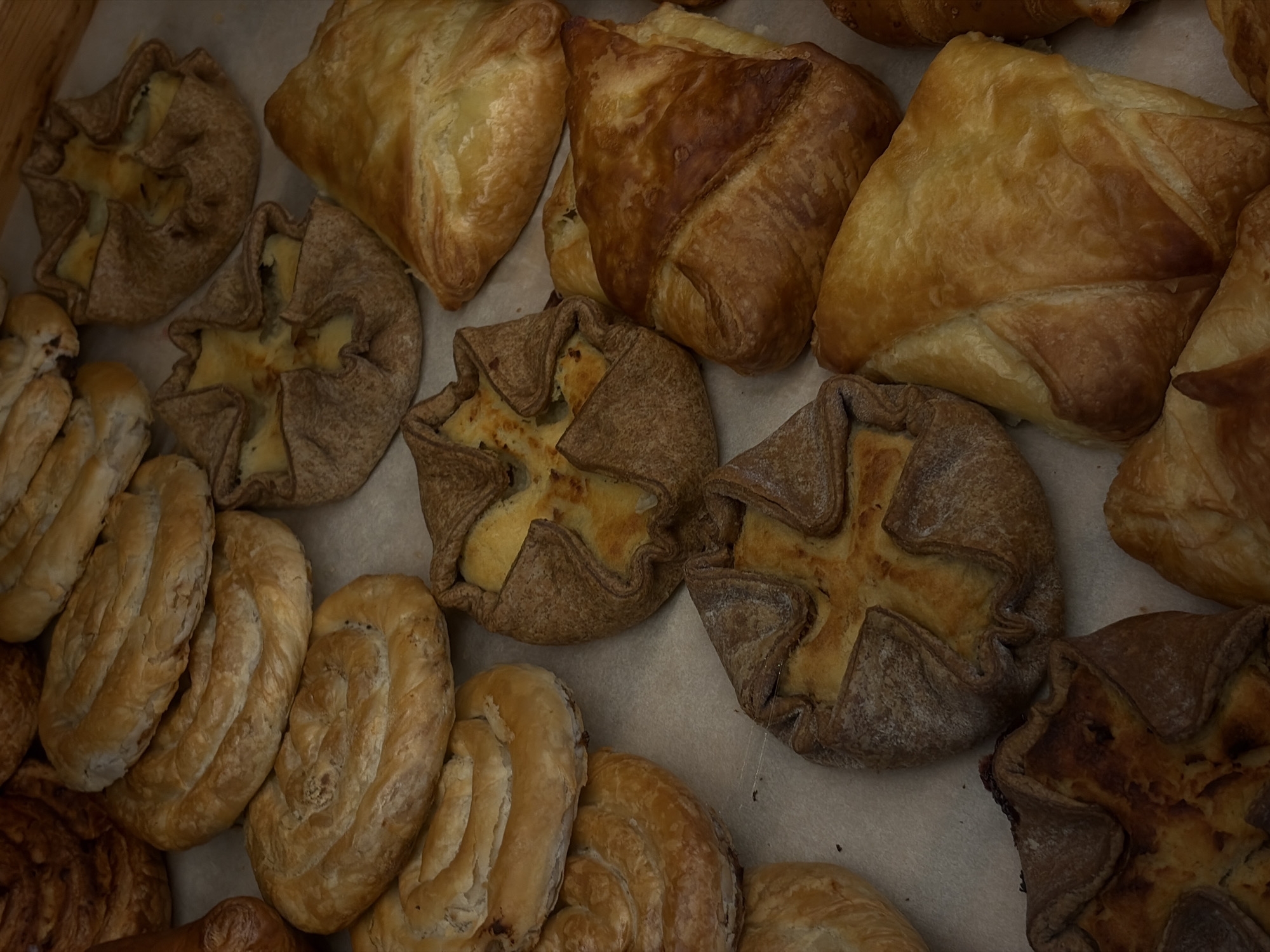 A close-up, top-down view shows an assortment of baked goods, likely pastries, arranged on a light-colored surface, possibly parchment paper. The pastries exhibit a variety of shapes and textures, including spiral-shaped buns, folded triangular pastries, and individual tart-like items with visible fillings. The colors range from golden brown to a deeper, slightly charred brown, indicating they are freshly baked. There are no people or discernible actions in the scene. The lighting is even, highlighting the details of the baked goods. This scene is likely from a bakery or market stall in Saint Petersburg, Russia, given the commonality of such pastries in Russian cuisine.