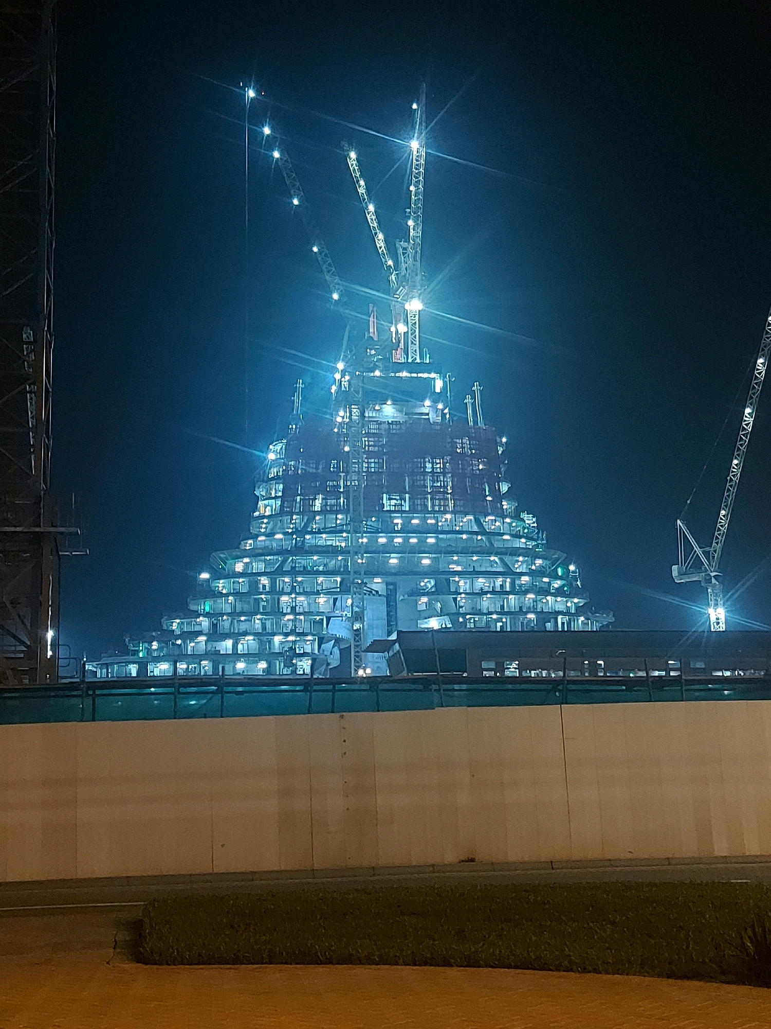 An exterior, full, horizontal, night-time shot captures a large, partially constructed building in an urban setting. The building has a unique, stepped, pyramid-like shape and is illuminated by numerous lights from within its many windows. Construction cranes are visible surrounding the structure, with one prominent crane on the right side of the frame. In the foreground, a low concrete wall with a green barrier on top runs horizontally across the image, separating the viewer from the construction site. Beyond the wall, a strip of green grass and a paved surface are visible. The sky is dark, suggesting it is nighttime.