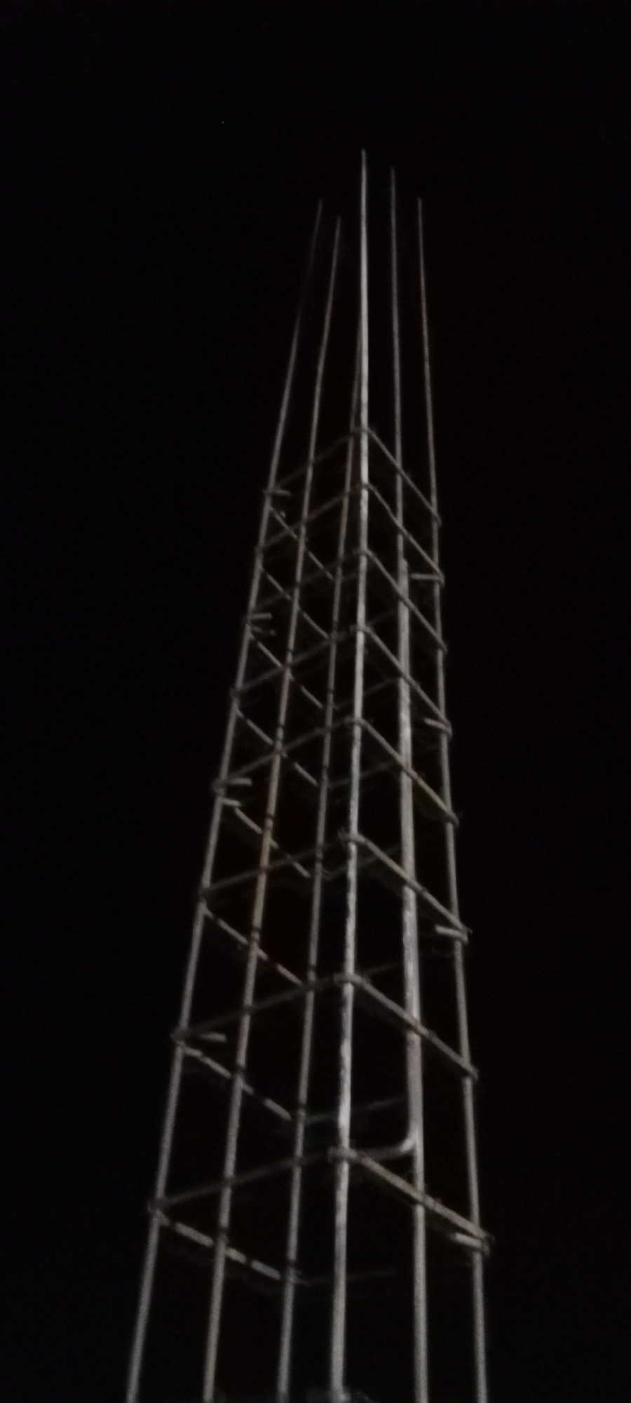 A tall, conical structure constructed from rebar is visible against a dark, possibly night sky. The rebar appears to be bundled and tied together to form a lattice-like frame that tapers to a point at the top. The structure is the primary subject of the image, dominating the frame. A single, faint point of light, possibly a star or distant light source, is visible in the upper left quadrant of the image against the black background. The image does not provide any specific details about the city of Soko, Indonesia, or any people, actions, or interactions. The scene is entirely focused on the rebar structure and its stark silhouette against the dark sky.