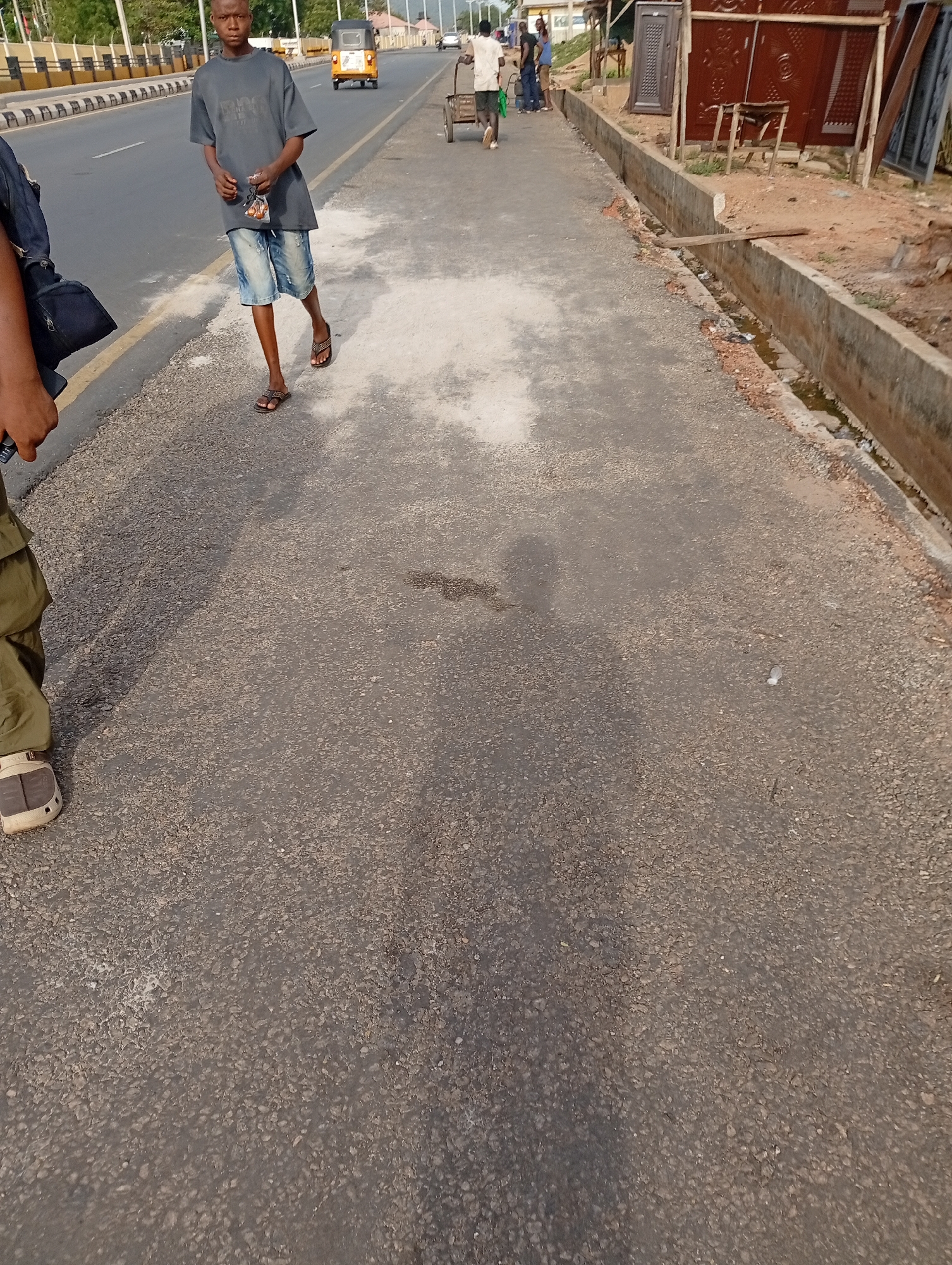 The image depicts a scene in Zango-ndaji, Nigeria, featuring a paved road and a sidewalk. On the sidewalk, a young man in a gray t-shirt, denim shorts, and sandals walks toward the camera, holding a small plastic bag. Behind him, another person walks away from the camera, pushing a cart. Further down the road, there is a yellow three-wheeled vehicle and other individuals. A concrete gutter runs along the edge of the sidewalk, near a building or structure with red doors. Part of another person is visible in the bottom left of the image. The ground shows dark shadows, possibly cast by the individuals in the scene.