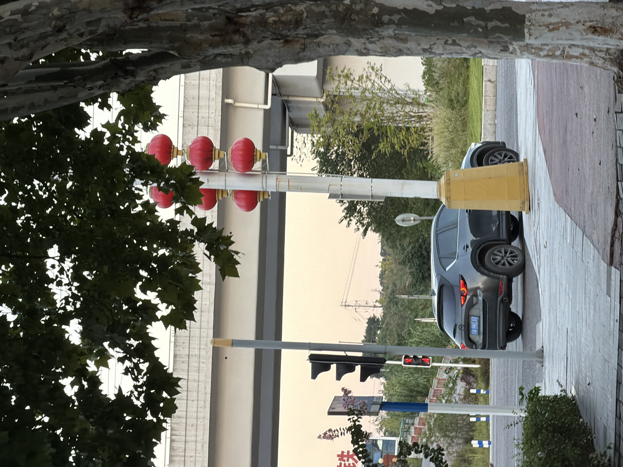 In Jiangjin, China, a dark gray SUV is parked on a sidewalk next to a road. The SUV has a license plate with Chinese characters and numbers. A traffic light is red and positioned above the vehicle. Red lanterns are hanging from a pole on the right side of the image, near the SUV. Trees and vegetation are visible in the background, and a concrete overpass is overhead. The scene appears to be an urban environment with street infrastructure.