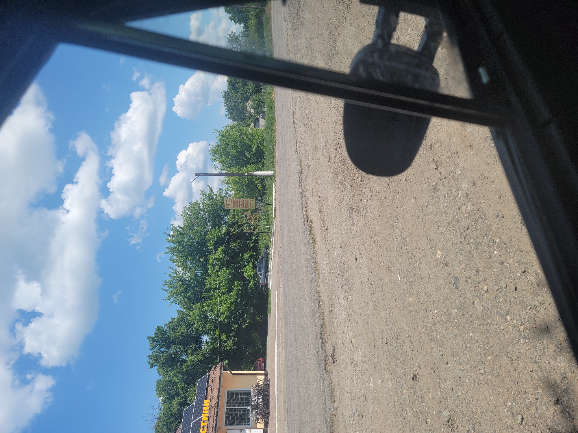 The image depicts a sunny day in Pereshchepyne, Ukraine, viewed from the perspective of someone inside a vehicle. In the foreground, a gravel road with some patches of wear is visible. To the left, a yellow building with solar panels on its roof is situated. The building has a storefront with a window displaying items and several bicycles parked in front of it. Beyond the building and road, a dense line of green trees forms a backdrop. A grey sedan is parked near the building, and further to the right, a white pole with signage is present. The sky is a vibrant blue, dotted with fluffy white clouds. A reflection of the vehicle's side mirror is visible in the lower right portion of the image, obscuring part of the scene.