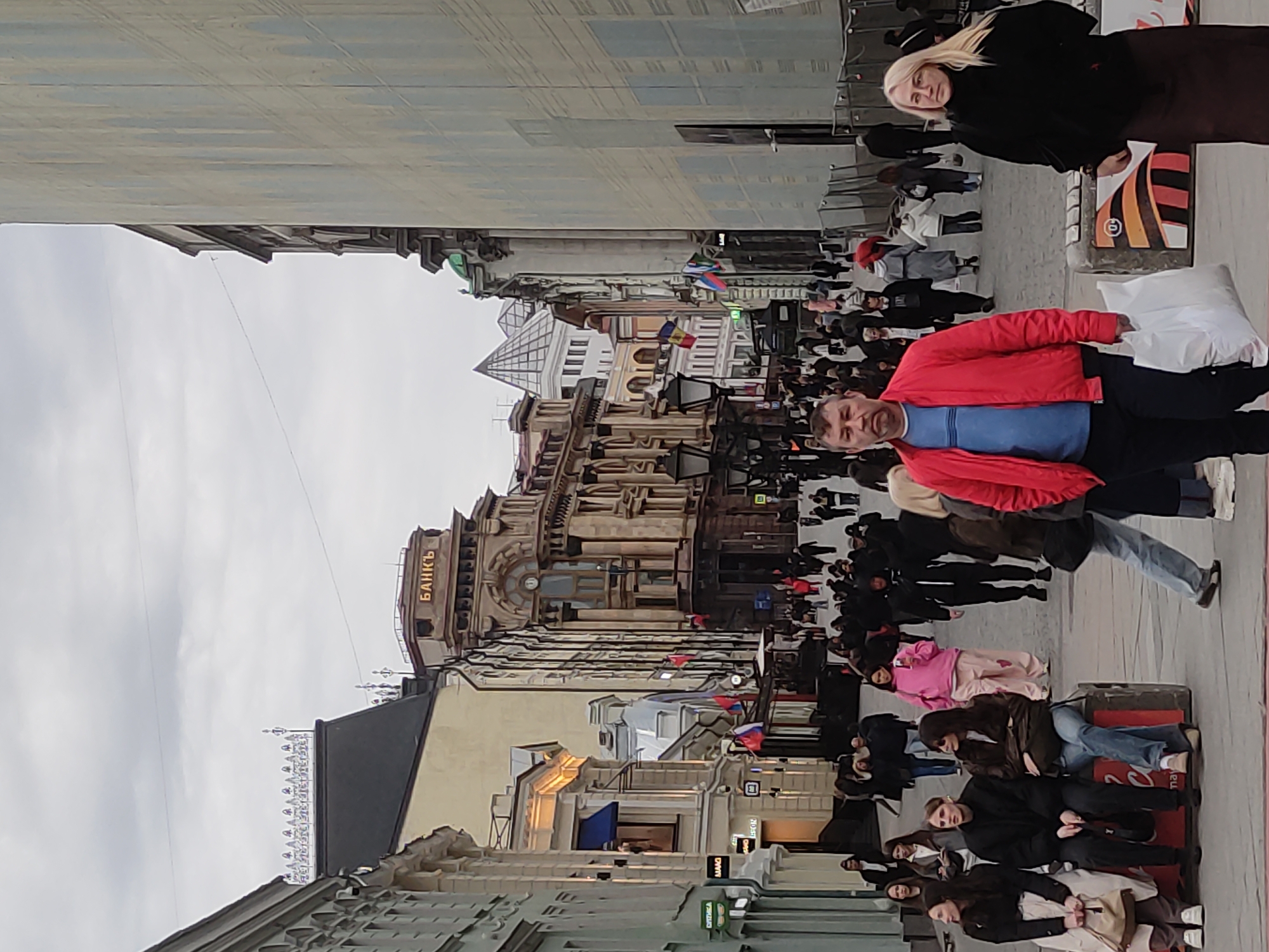 The image depicts a bustling street scene in Moscow, Russia. People are walking along the sidewalk, some carrying shopping bags. One individual is wearing a bright red jacket and carrying a white bag. Buildings line the street, with one prominently displaying the word "БАНКЪ" in Cyrillic letters, indicating a bank. Russian flags are visible on some buildings. The sky is overcast.