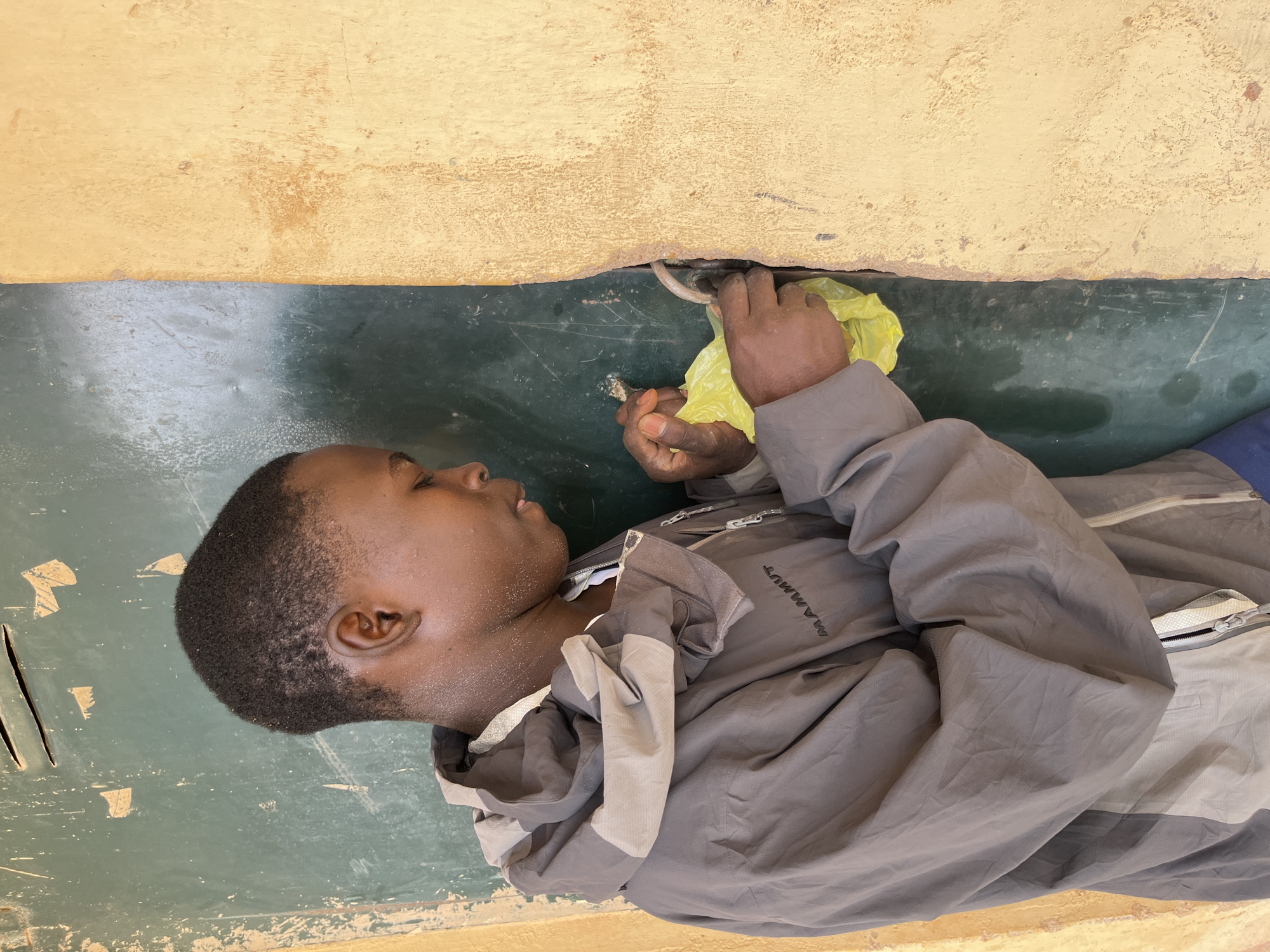A young person wearing a grey and beige hooded jacket with the logo "MAMMUT" is standing next to a weathered, green door frame. Their right hand is holding a bright yellow plastic bag and is reaching into a gap between the door and the frame. The person's head is tilted down as they focus on the action. The environment appears to be outdoors, with a textured, light-colored wall adjacent to the door. The overall scene suggests an activity taking place in a rustic or possibly a less-maintained urban setting.