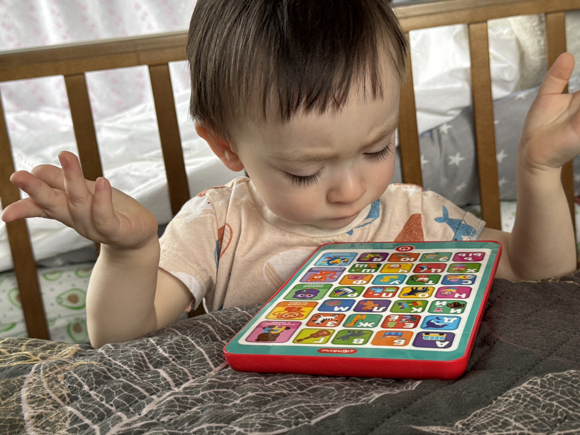 A toddler with short brown hair, wearing a patterned light-colored t-shirt, sits in front of an educational tablet featuring Russian letters. The child has a confused expression, with eyebrows furrowed, looking down at the tablet. Their hands are raised in a gesture of uncertainty. The tablet is red and has a grid of squares, each with a letter and a corresponding image. The background consists of the wooden bars of a crib and a patterned blanket. The location is likely a home setting. The image does not include specific details identifying it as Izhevsk, Russia, or any other city.