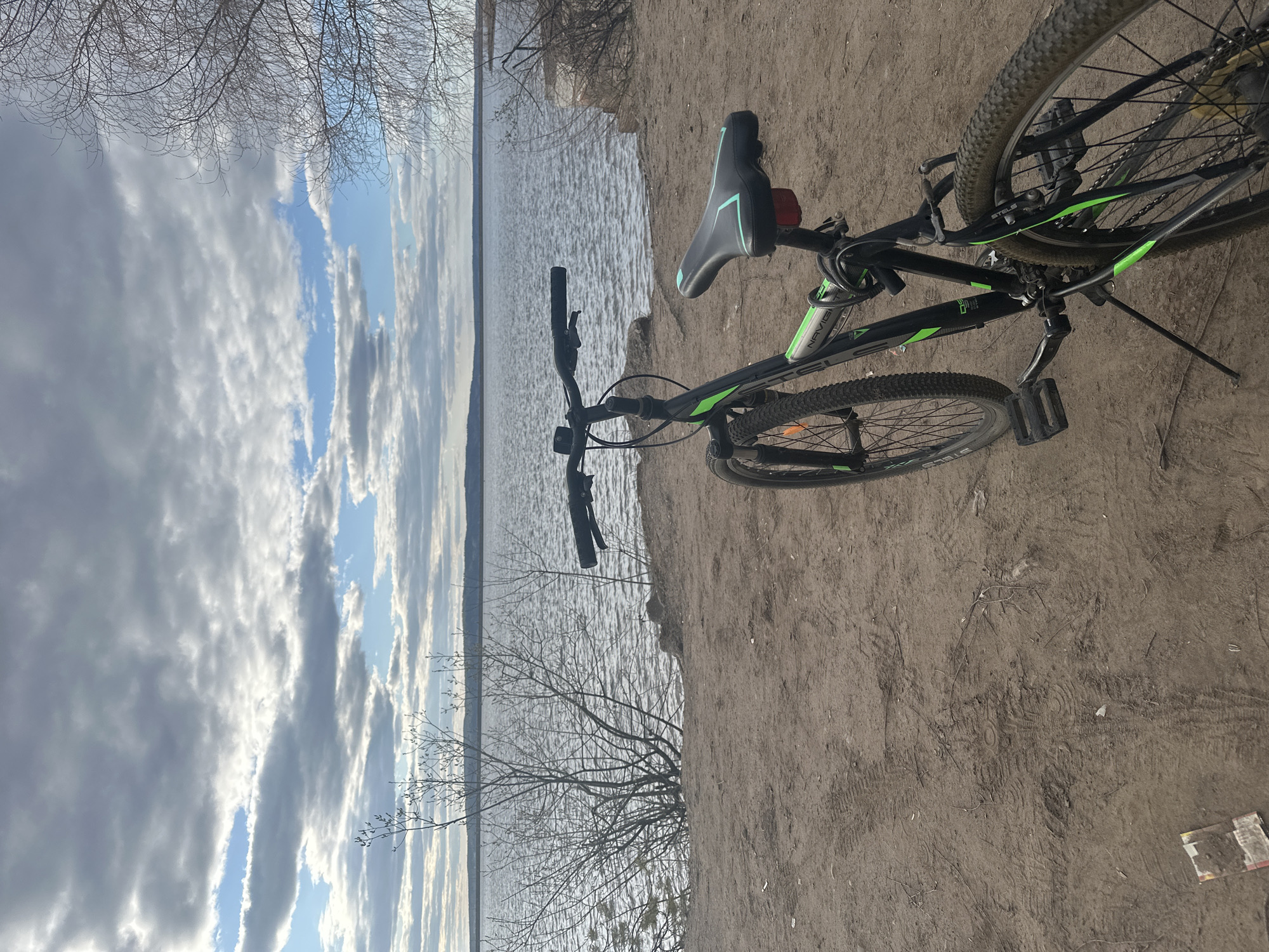 The image shows a bicycle parked on a sandy bank next to a body of water. The bicycle is black with green accents and is resting on its kickstand. The water is calm with some ripples and reflects the sky, which is filled with clouds. The sandy bank appears to be dirt-like sand with dried debris. The location is likely near a body of water, possibly in the city of Chaikovsky, Russia, given the prompt's context. No people are visible in the image. The bicycle is stationary. The image captures a moment of rest or pause during an activity involving the bicycle near the water.