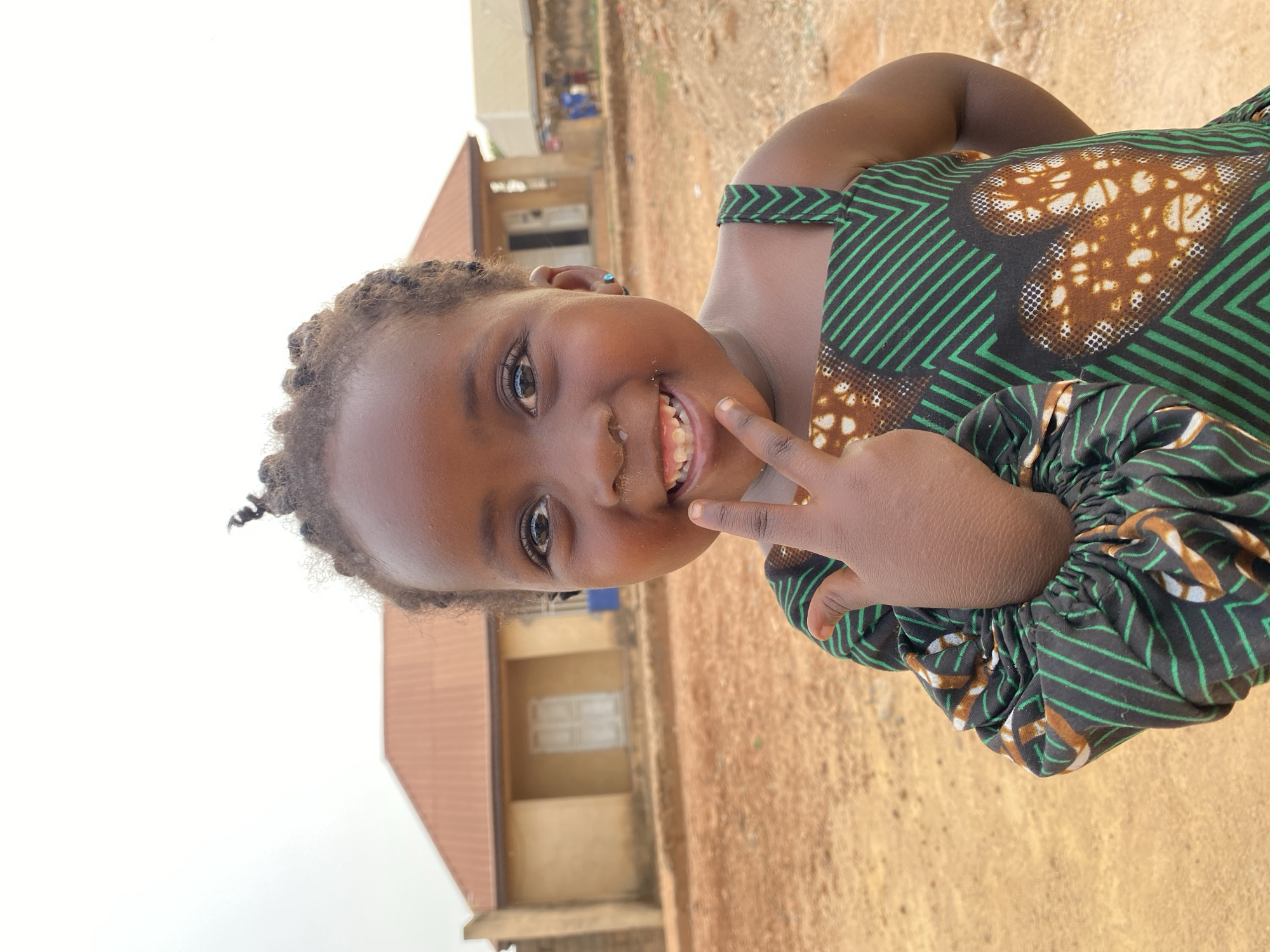 A young girl is smiling and holding up two fingers in a peace sign. She has dark skin and is wearing a green and brown patterned dress. Her hair is braided. The background shows a building with a red roof and a dirt ground. This image appears to be taken in Kuru Dungus, Nigeria.