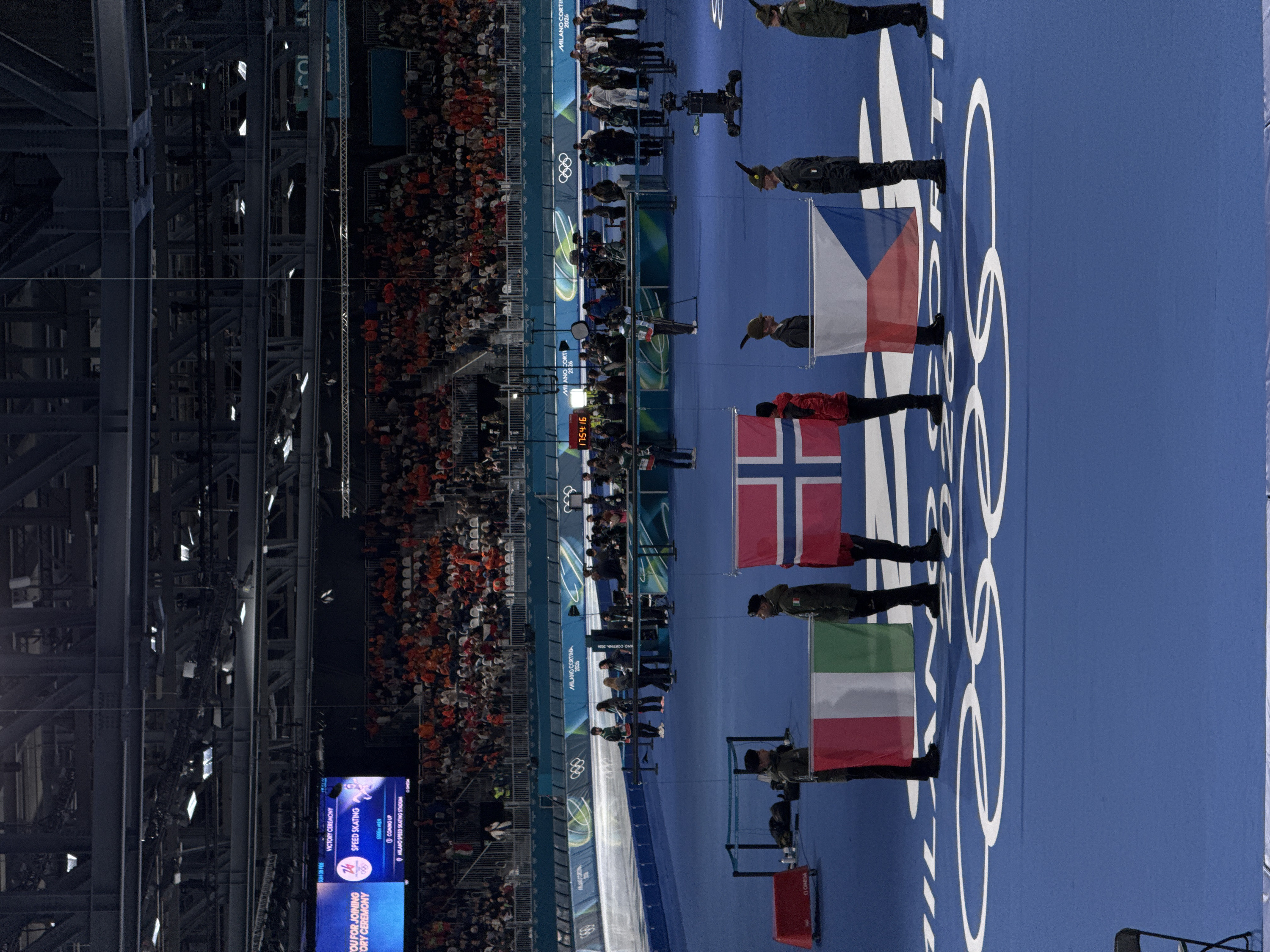 The image shows a speed skating victory ceremony at the Milano Speed Skating Stadium for the 2026 Milano Cortina Winter Olympics. Guards of honor are holding flags of Italy, Norway, and the Czech Republic. The venue is full of spectators. A large screen displays "Victory Ceremony," "Speed Skating," and "6000m Men," along with the time "17:54:16." Olympic rings are visible on the floor.