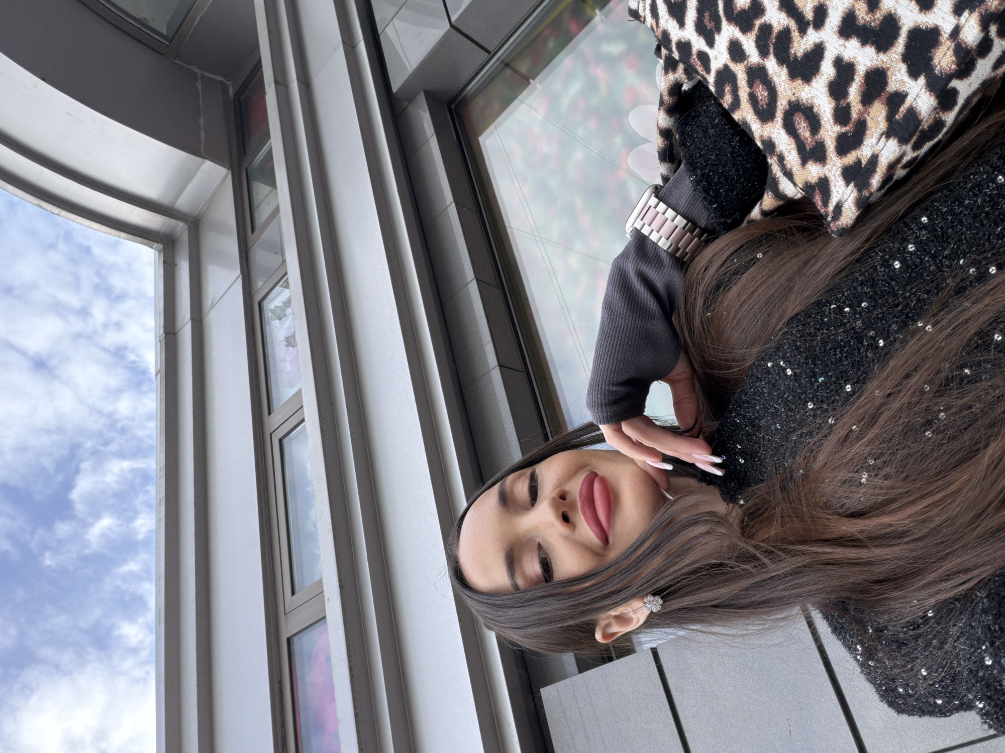 A woman is positioned outdoors, near a building in Saint Petersburg, Russia. She has long brown hair, visible earrings, and wears a black long-sleeved top embellished with sequins, as well as a jacket/blanket with a leopard print. Her right hand is slightly raised near her face, displaying long nails. She is wearing a watch on her left wrist. The background includes a modern building with vertical architectural elements and windows reflecting outdoor light, possibly indicating the sky or other buildings. The sky visible through the windows is partially obscured by clouds.