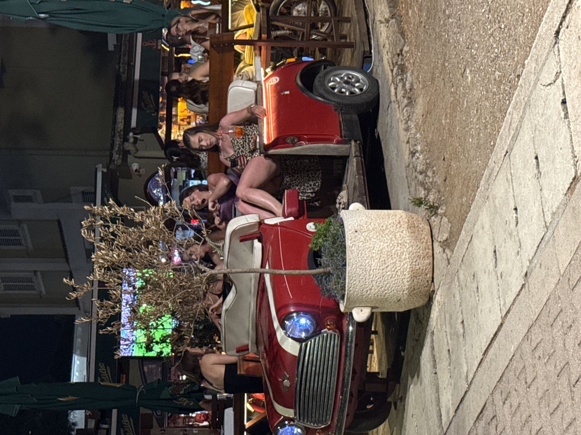 The image depicts an outdoor scene at night, likely in the city of Budva, Montenegro, given the aesthetic. In the foreground, a red, open-top Mini Cooper serves as a seating area for several young women. One woman, wearing a leopard-print dress, is seated in the driver's position and holds a drink and a phone. Another woman next to her is yawning. Behind them, more people are visible at tables, with one person appearing to watch a sports game on a screen in the background. The Mini Cooper is positioned on a paved area next to a sidewalk, with a large potted plant containing a small tree and purple flowers to its left. The overall atmosphere suggests a lively outdoor dining or bar setting.
