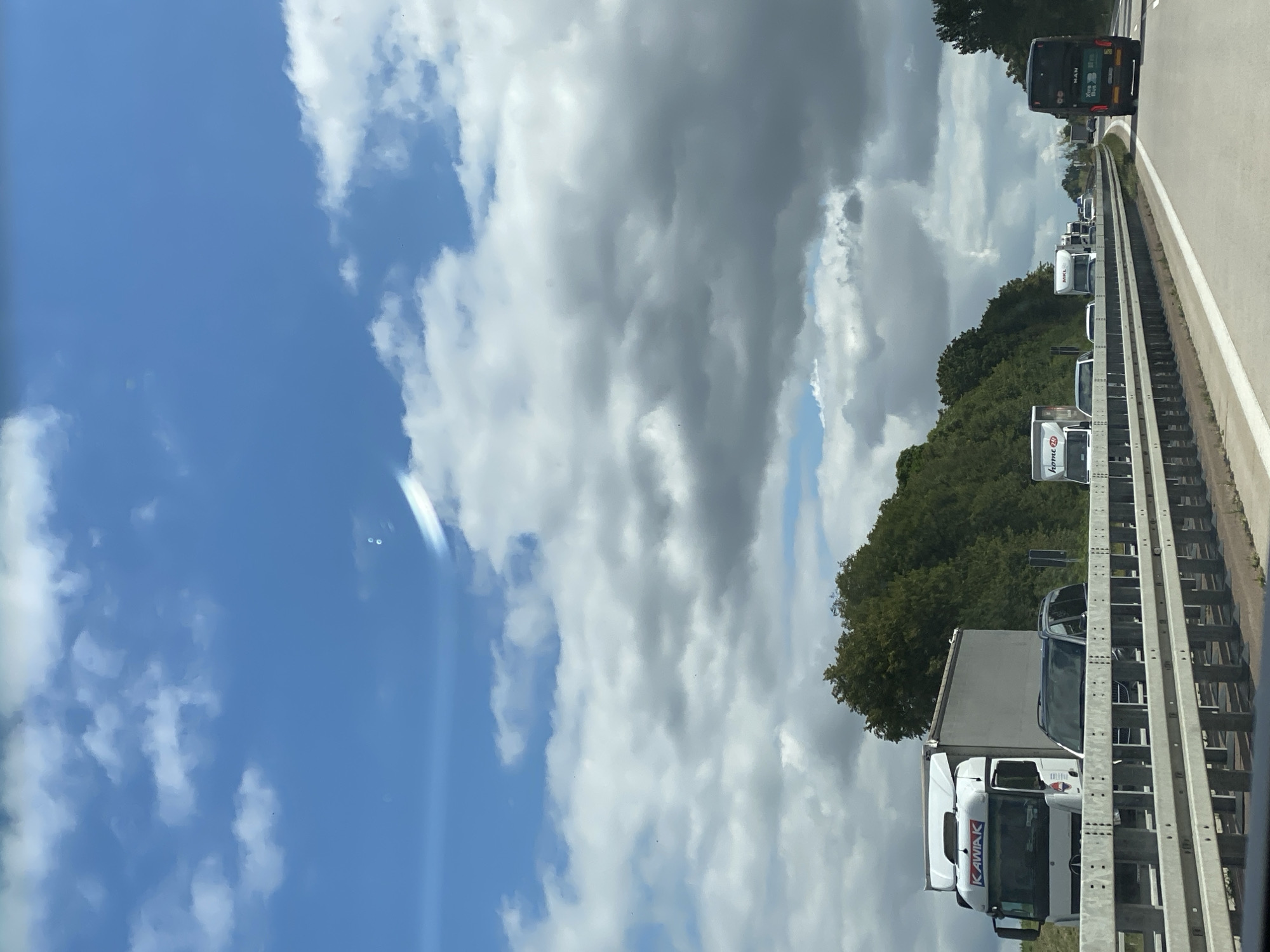 The image captures a highway scene in the vicinity of Nossen, Germany. The perspective is from within a vehicle traveling on the highway. In the foreground, a concrete barrier with horizontal railings separates lanes of traffic. Several vehicles are visible on the highway, including white semi-trailer trucks with company logos such as "KAWIAK" and "home 24", a dark-colored SUV, and a black bus with "MAN" branding. The highway appears to be a multi-lane road. On the left side of the highway, a dense line of green trees forms a natural embankment. The sky above is a mix of blue sky and large, gray clouds, suggesting a partly cloudy day. The overall scene depicts moderate traffic flow on a European highway, surrounded by a wooded landscape.