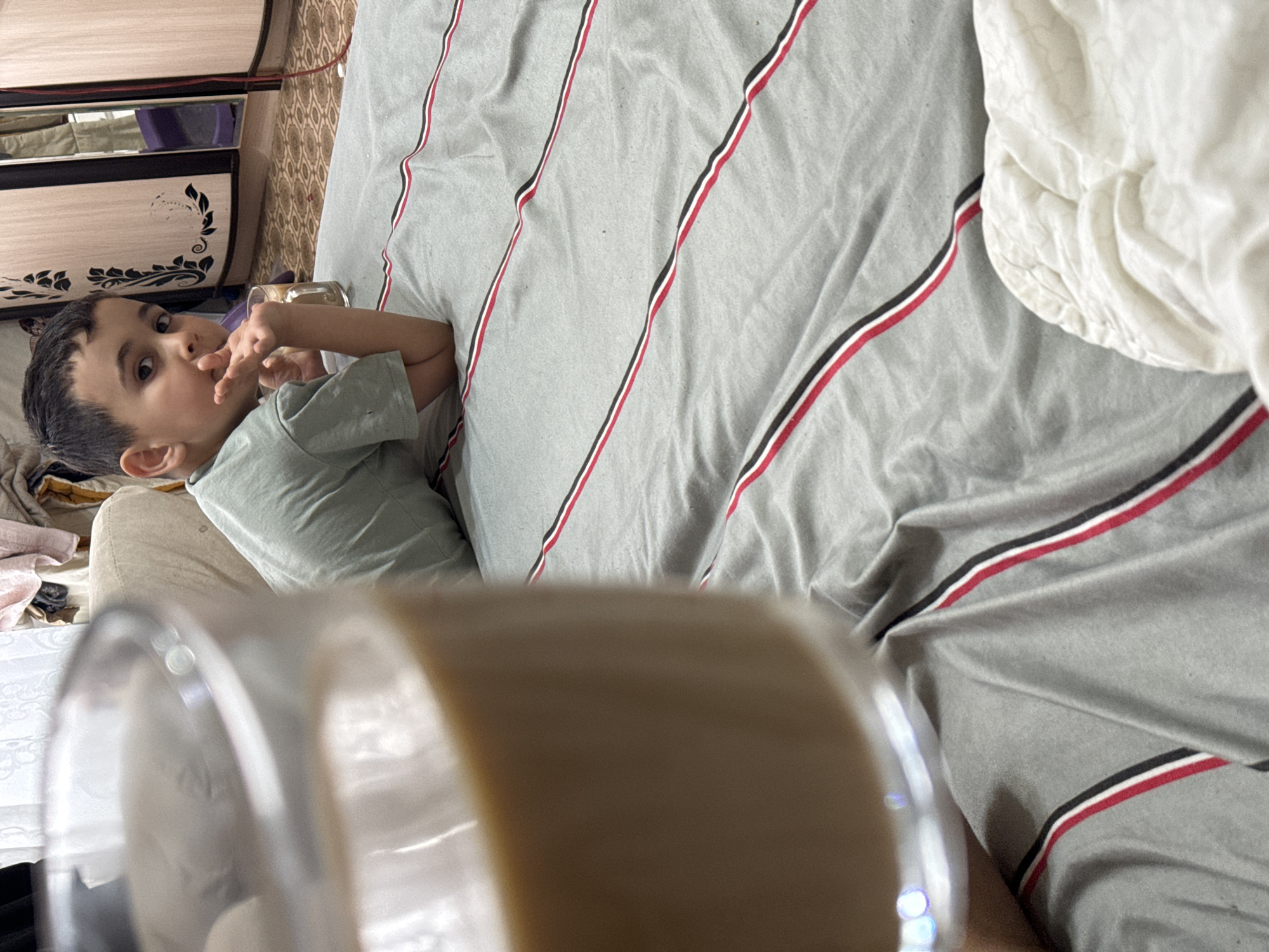 The scene captures a young boy lying on a bed covered with a gray striped duvet. The boy is wearing a light green t-shirt and is looking at the camera while holding his fingers up to his mouth. To the left of the frame, there is a piece of furniture with a black and white floral design. In the foreground, there is a clear glass partially filled with a light brown liquid, obscuring parts of the scene. Based on prior data association of the user, the geographical location is Chaikovsky, Russia.