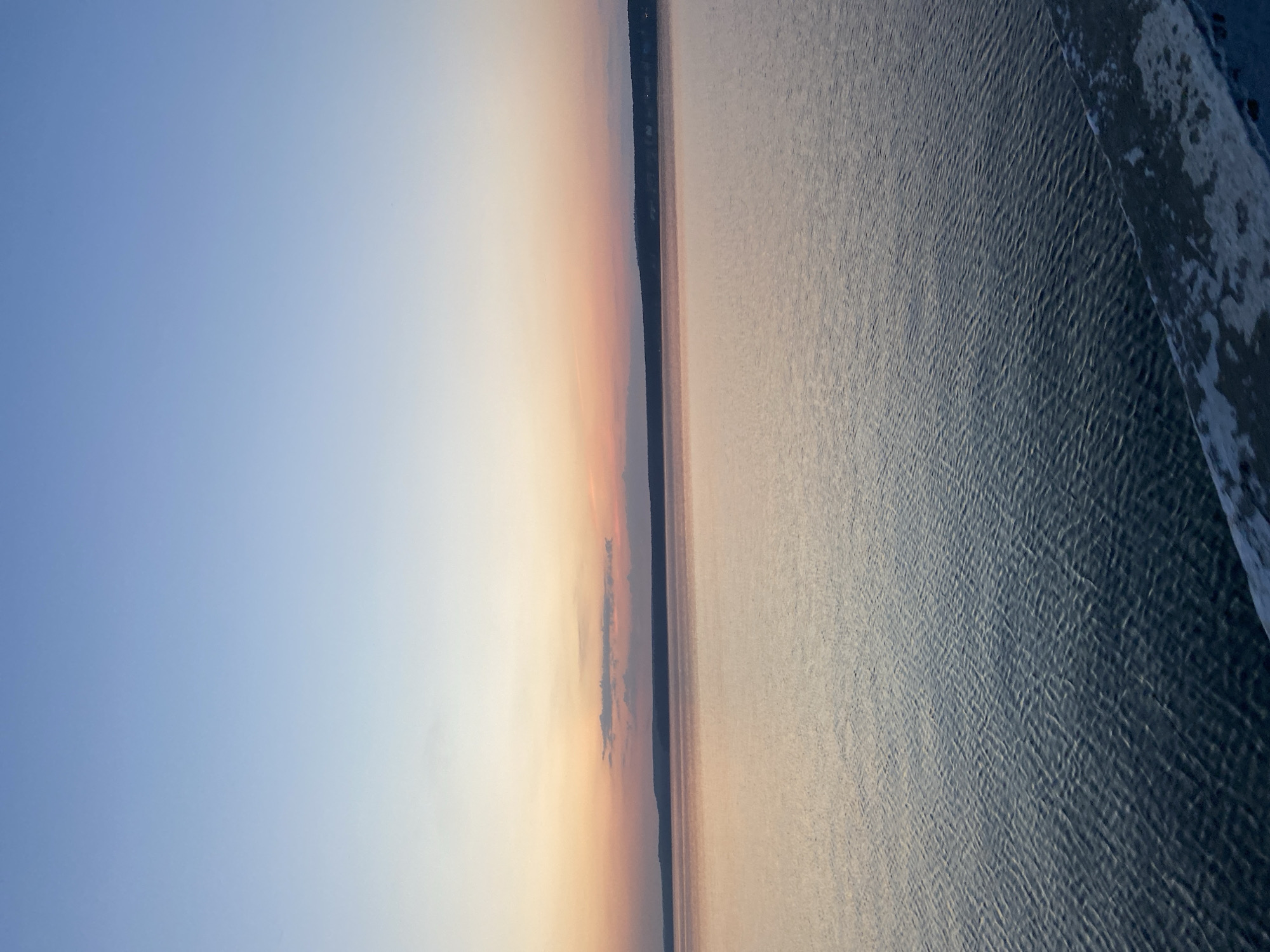 The image presents a view of the Votkinsk Reservoir in Votkinsk, Russia. The sky transitions from a pale blue to orange hues near the horizon, indicating sunrise or sunset. A dark, narrow strip of land is visible along the horizon, separating the sky from the water. The water surface exhibits ripples and variations in texture, reflecting the ambient light. Along the top edge of the frame, there is a strip of snow or ice along the shoreline.