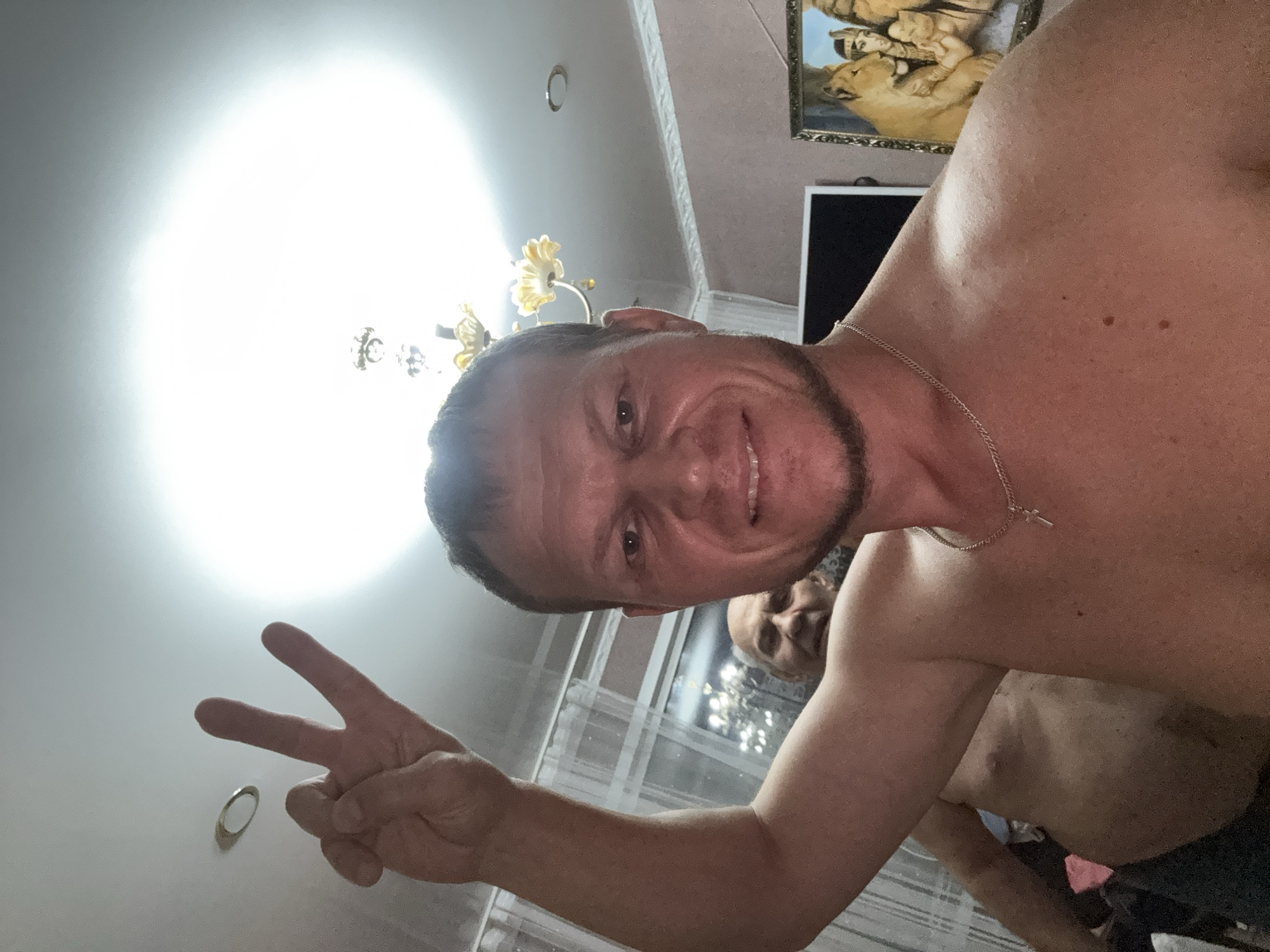 In the foreground, a shirtless man with short brown hair and stubble is taking a selfie. He is smiling and holding up his left hand in a peace sign. His right arm is raised in the air. He is wearing a silver chain with a cross pendant. In the background, another shirtless man with gray hair is visible. The room appears to be a living space with a chandelier on the ceiling, a painting of lions on the wall, and a television screen. The lighting is bright, suggesting an indoor setting, likely Glubokoe, Kazakhstan, though this cannot be confirmed by the image itself.