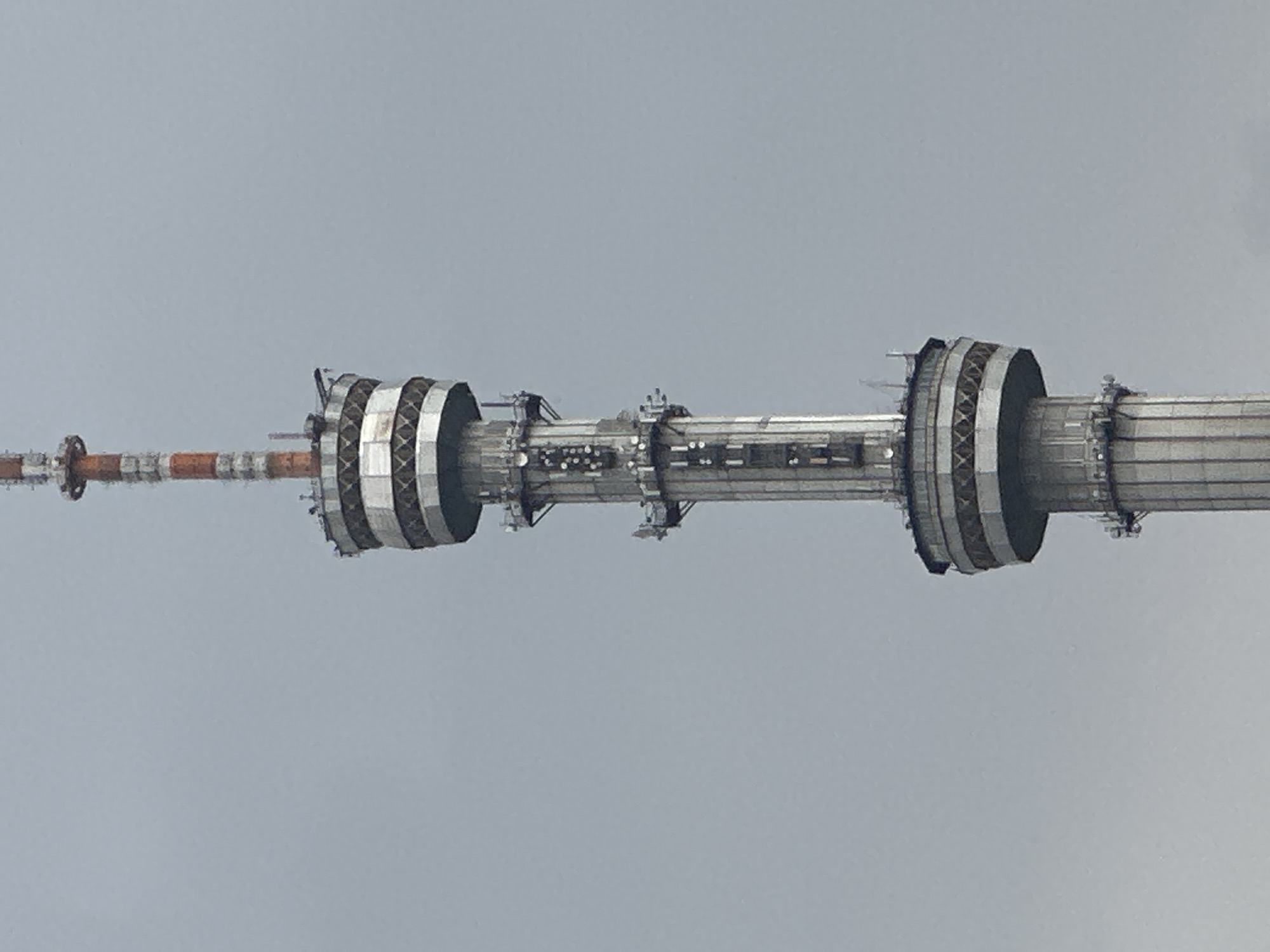 The image shows the upper section of the Almaty Television Tower, also known as the Kok-Tobe Tower, located in Almaty, Kazakhstan. The tower is a prominent structure with multiple platforms and antennas. The sky is overcast and grey. There are no people visible in the image. The tower is made of metal and concrete, and appears to be a functional telecommunications and broadcast tower.