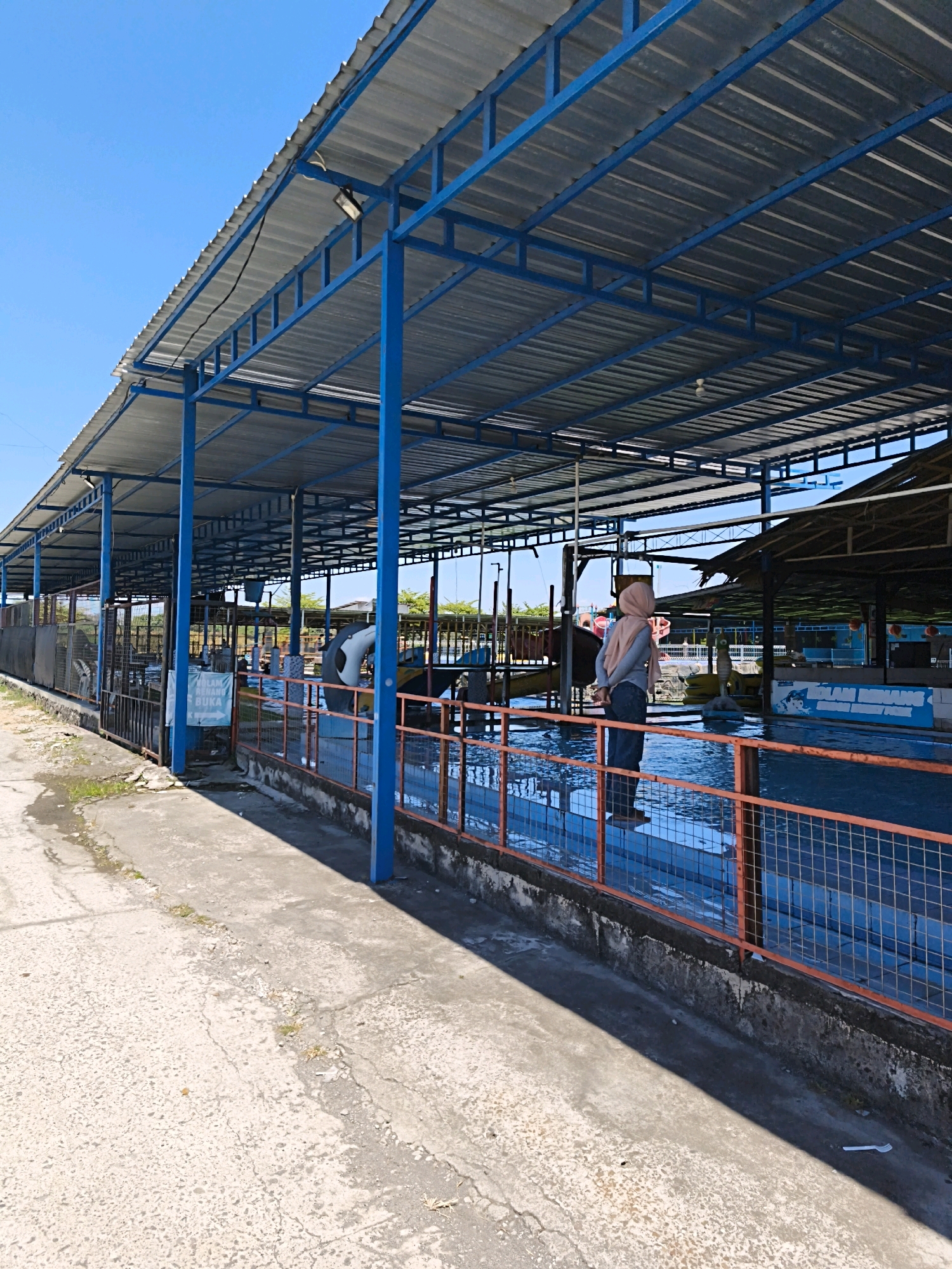 The image depicts an outdoor recreational facility, likely a water park or swimming pool area, under a bright blue sky. The foreground shows a cracked concrete pathway with some debris. A long, covered structure with a corrugated metal roof supported by blue metal beams runs along the side of the pathway. Orange metal railings with mesh infill line the edge of the water area.
In the water, a mannequin, possibly a decorative statue or part of an attraction, is visible. It appears to be a nude or partially clothed figure with a light skin tone. The water is a vibrant blue, suggesting it is a swimming pool. Various other structures, including what looks like a slide and other pool-related equipment, are visible in the background. A sign with Indonesian text is also present, though it is partially obscured. The overall scene suggests a warm, sunny day at a public water attraction in Desa Sindon, Indonesia.